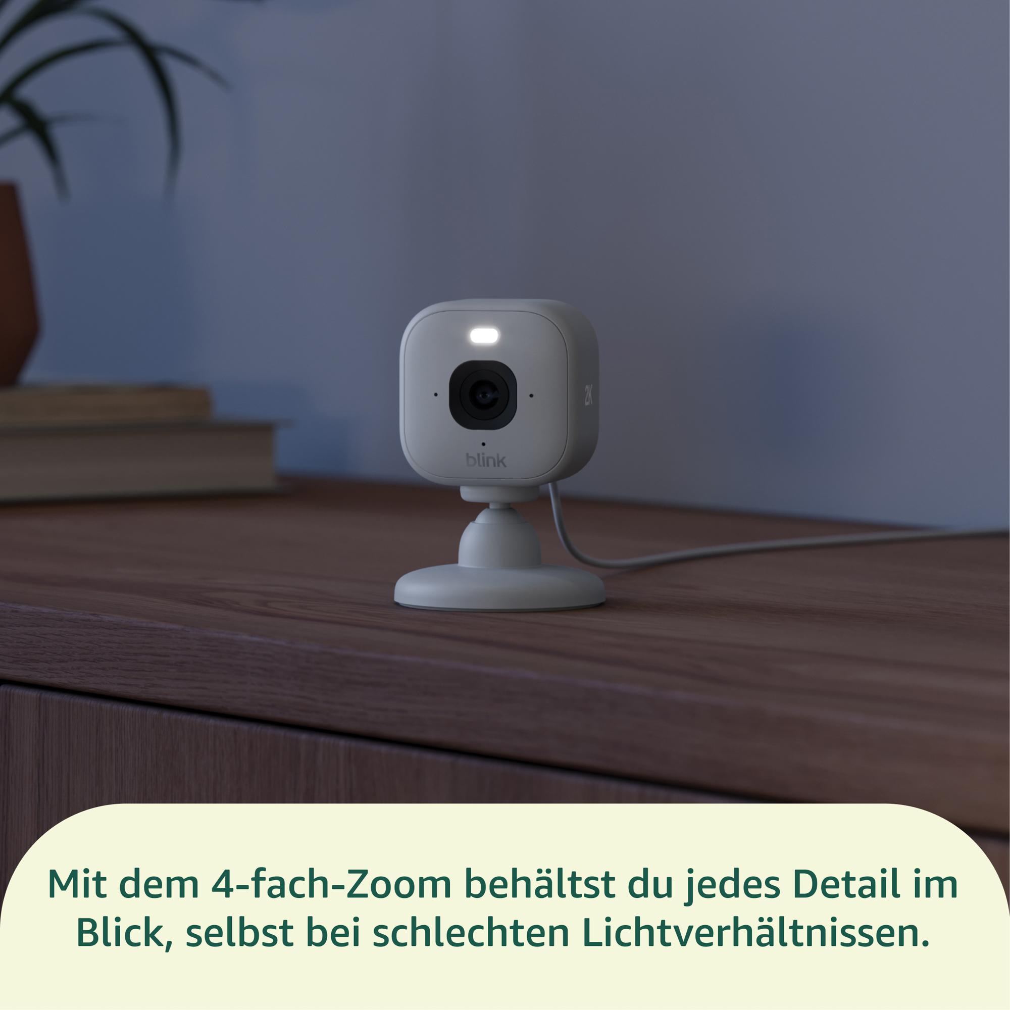 Eine weiße Blink-Überwachungskamera auf einem Ständer auf einer Holzoberfläche. Eine Pflanze und Bücher sind im Hintergrund.