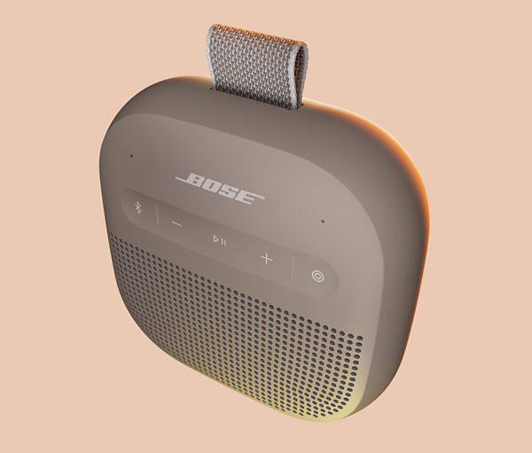 Ein beigefarbener Bose-Lautsprecher mit Stoffgriff und Steuertasten. Er steht auf einem pfirsichfarbenen Hintergrund. Der Lautsprecher hat eine Mesh-Front.