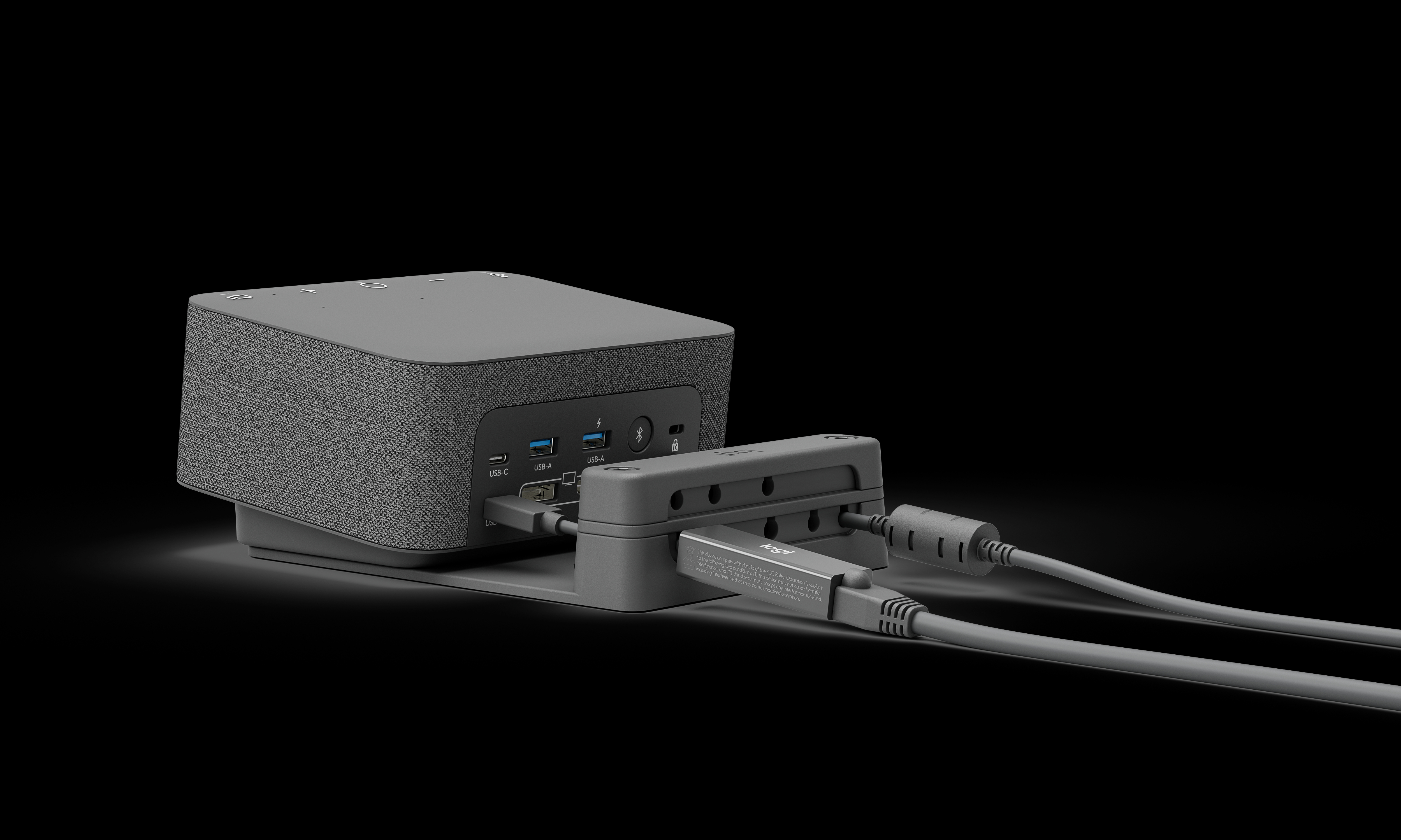 Ein grauer Lautsprecher mit USB-Anschlüssen und angeschlossenen Kabeln, auf einem Ständer vor schwarzem Hintergrund.