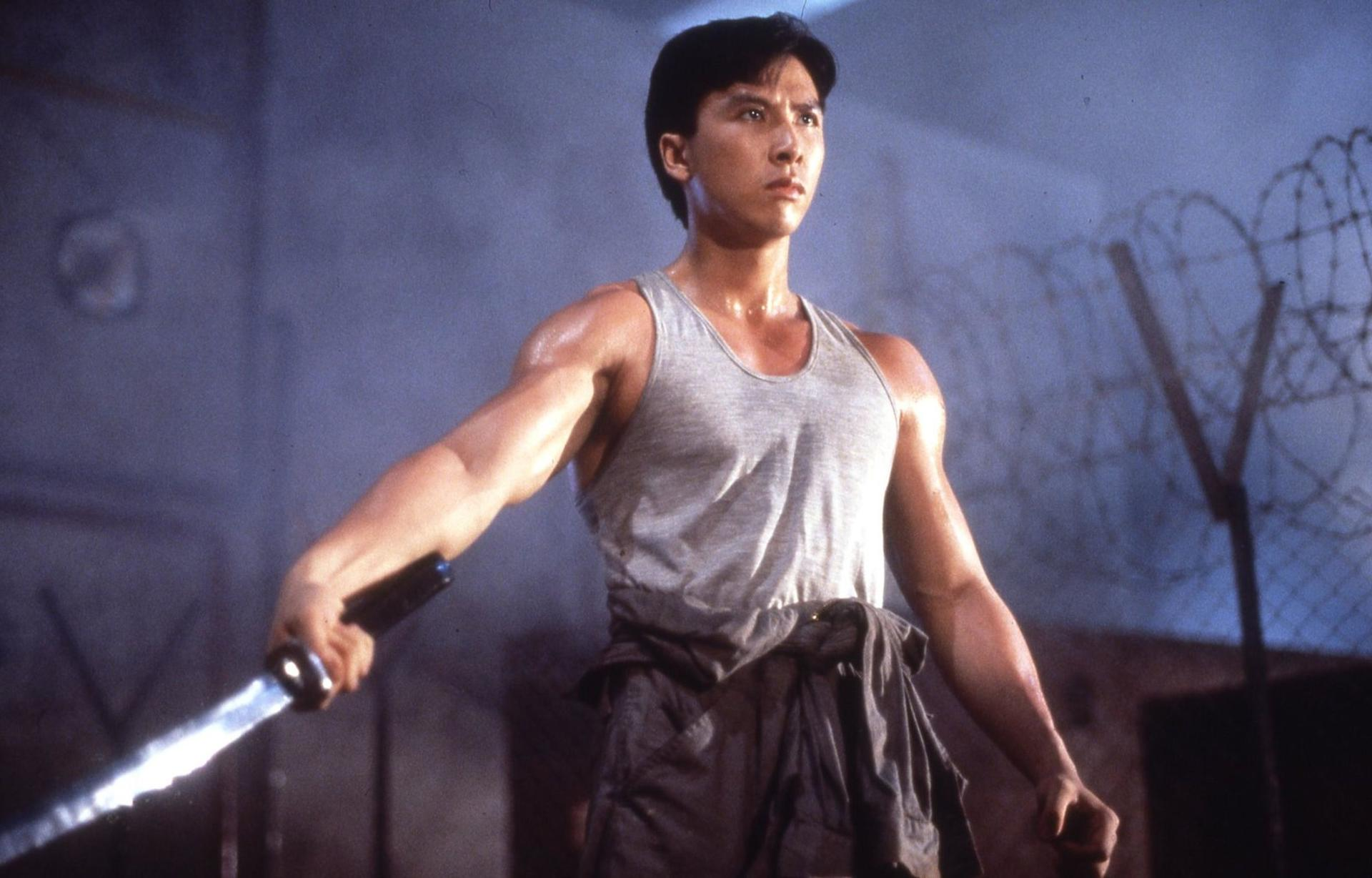 Ein Mann in einem grauen Tanktop hält ein leuchtendes Schwert. Er steht vor einem Stacheldrahtzaun. Der Hintergrund ist ein dunkler Raum.