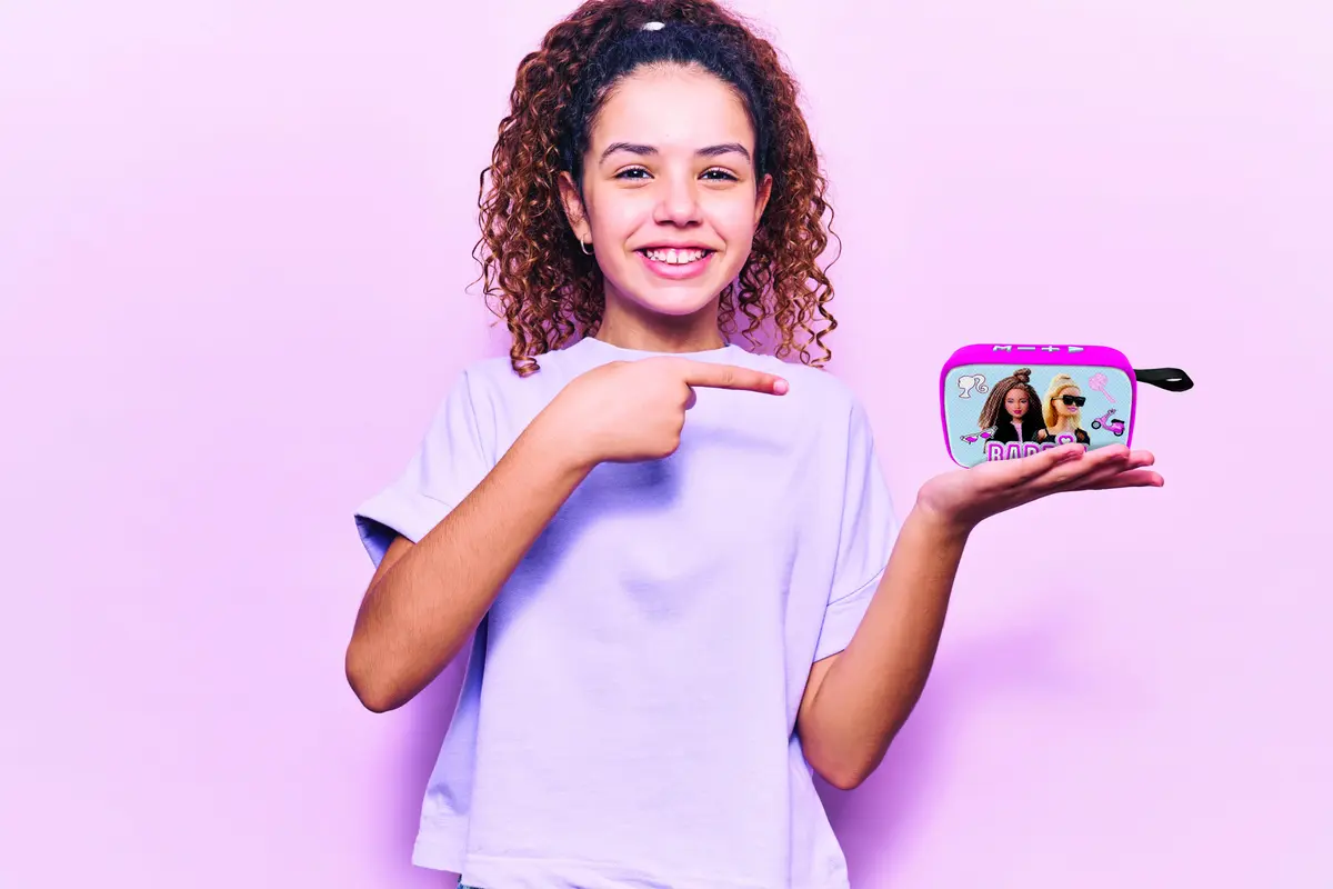 Una niña sonriente señala un altavoz rosa con imágenes de Barbie. Lleva una camisa blanca sobre un fondo rosa.