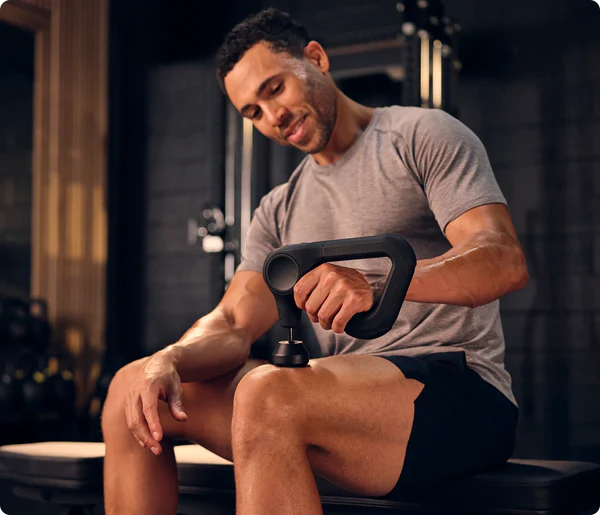 Ein Mann in einem Fitnessstudio benutzt eine Massagepistole an seinem Knie. Er trägt ein graues Hemd und schwarze Shorts. Im Hintergrund sind Fitnessgeräte zu sehen.