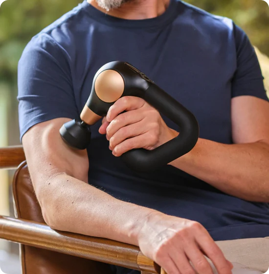 Ein Mann massiert seinen Arm mit einer schwarz-goldenen Massagepistole. Er trägt ein blaues Hemd und sitzt auf einem braunen Stuhl.