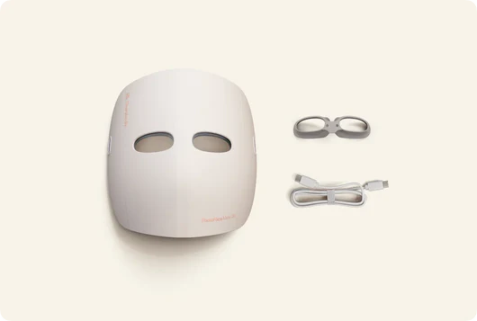 Eine weiße LED-Gesichtsmaske, eine Brille und ein Ladekabel liegen auf einem hellbeigen Hintergrund. Die Maske hat Augenlöcher.
