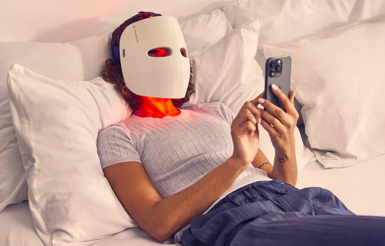 Person mit Lichttherapiemaske, benutzt Handy im Bett. Rotes Licht strahlt aus der Maske.