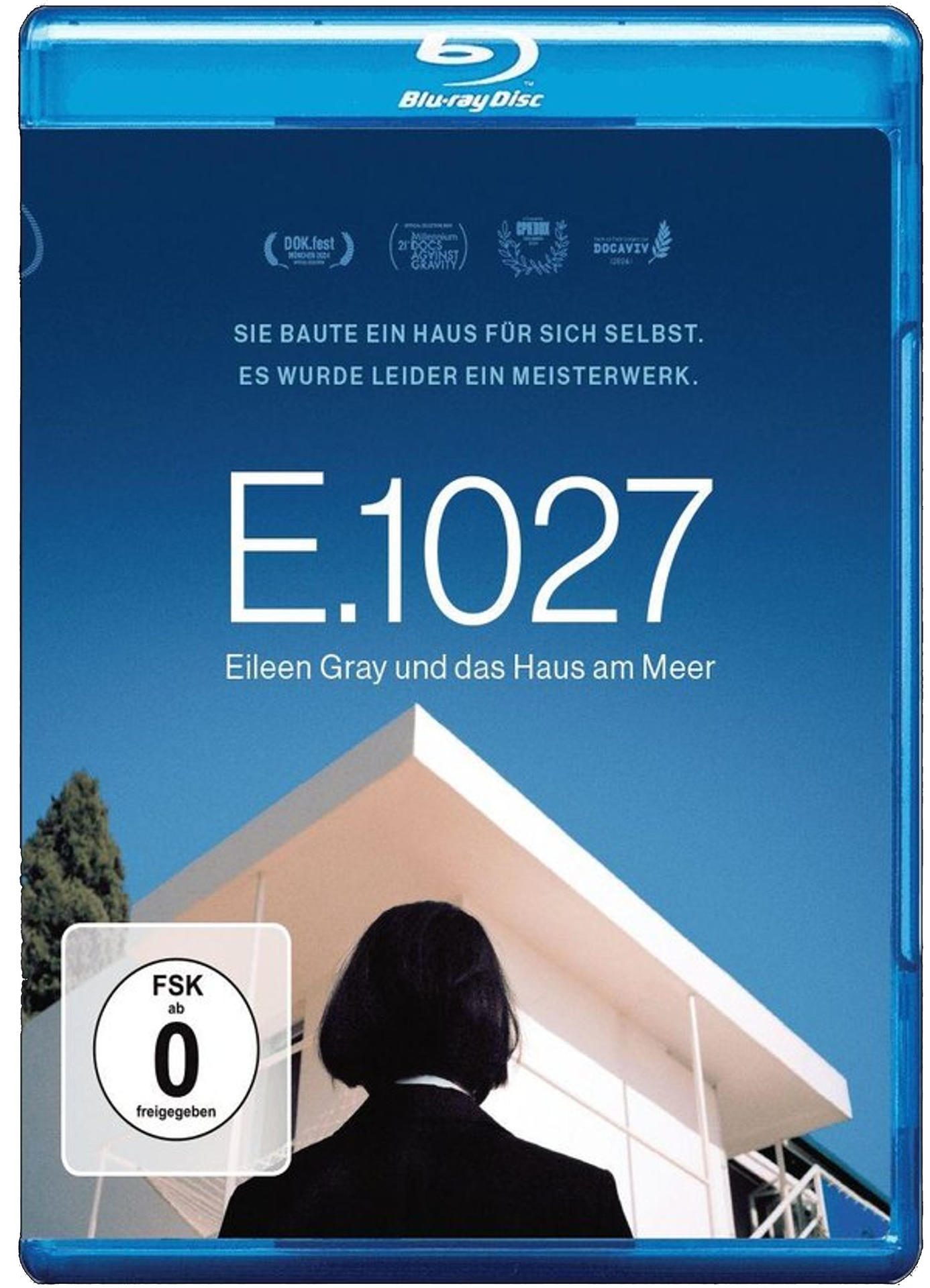 Blu-ray-Cover. Titel: E.1027. Eine Frau in dunklem Anzug steht vor einem modernen weißen Gebäude. Der Himmel ist blau.