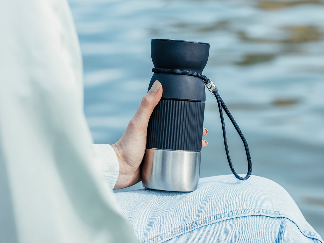 Eine Person hält einen dunkelblauen und silbernen Reisebecher. Die Person trägt eine hellblaue Jeans und einen weißen Pullover. Hintergrund ist Wasser.