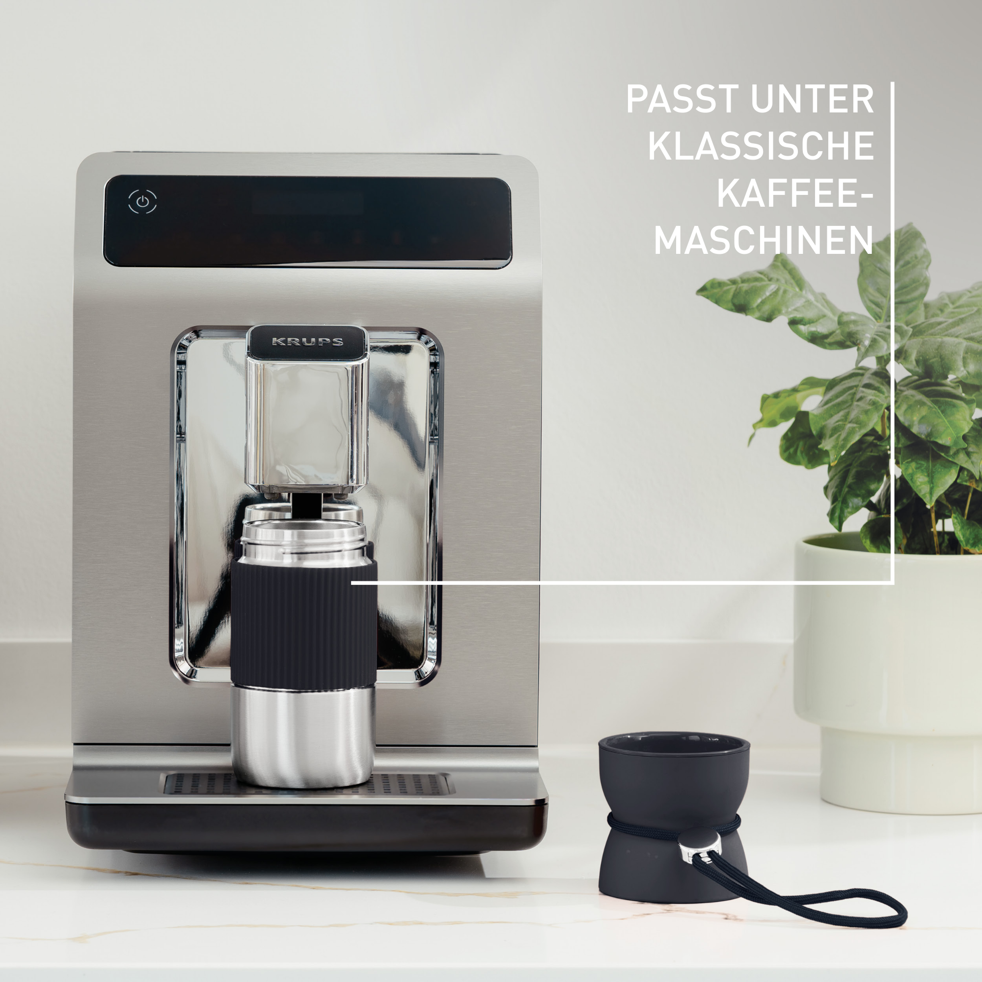Eine silber-schwarze Krups-Kaffeemaschine mit schwarzer Tasse. Rechts eine Pflanze im weißen Topf.
