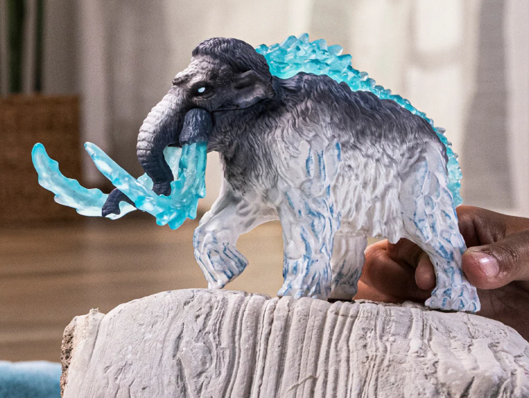 Ein Spielzeugmammut mit blauen Eiseffekten steht auf einem Stein. Das Mammut ist grau und weiß, mit einer Hand, die es hält.
