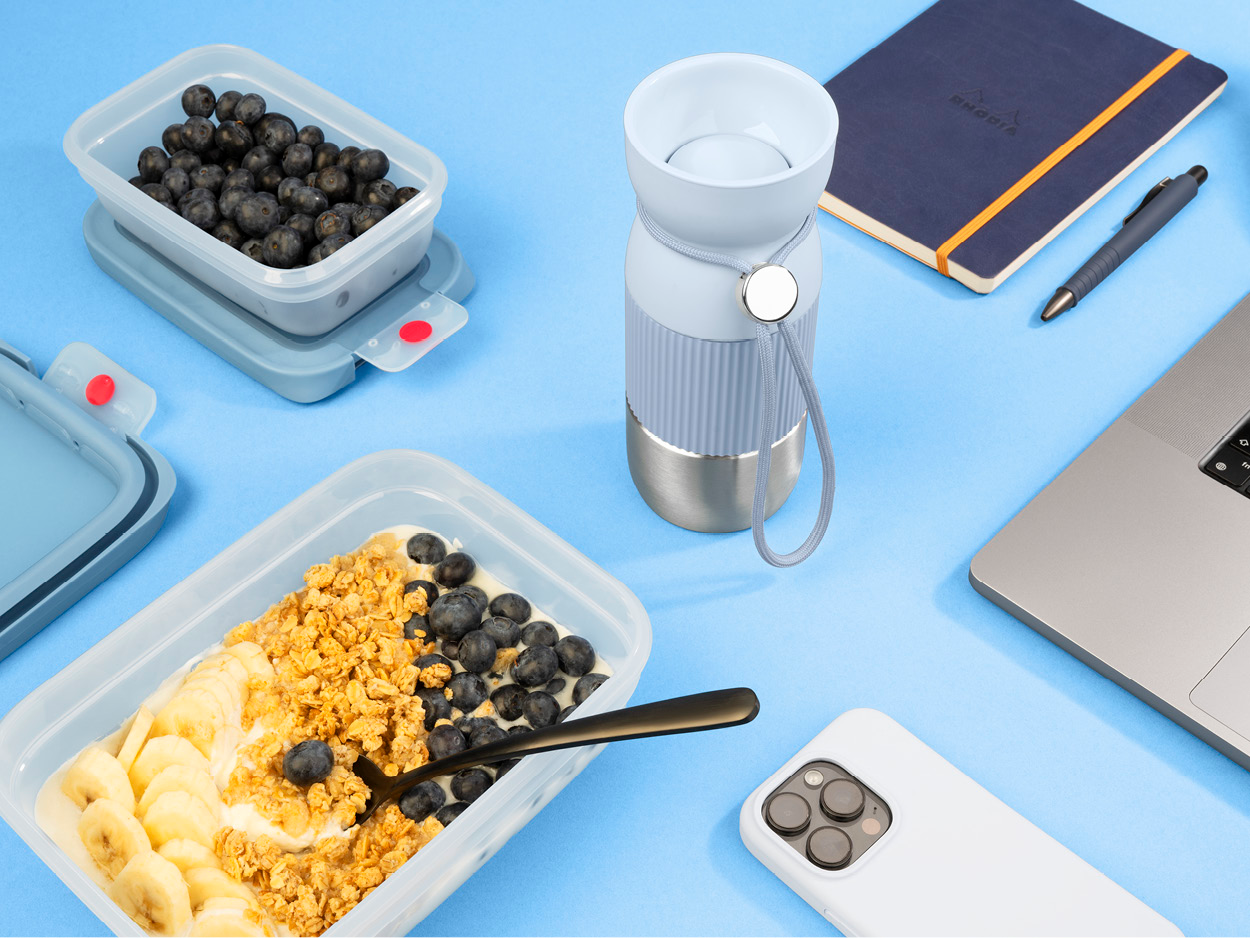 Blaubeeren in einem Behälter, Joghurt mit Müsli und Bananenscheiben, eine Wasserflasche, Notizbuch, Stift, Laptop und Telefon auf Blau.