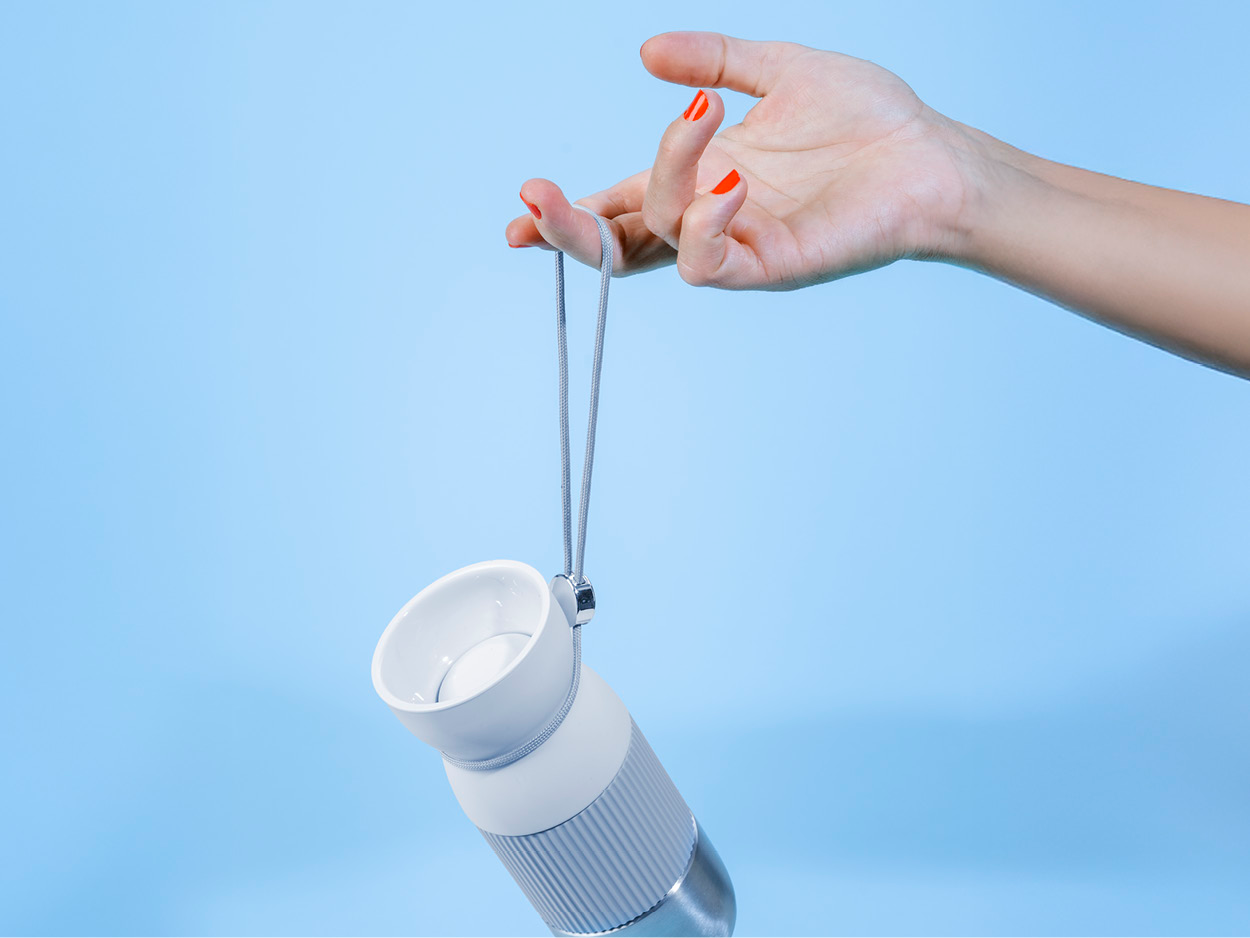 Eine Hand mit rotem Nagellack hält eine weiß-graue Wasserflasche mit grauem Riemen vor blauem Hintergrund.