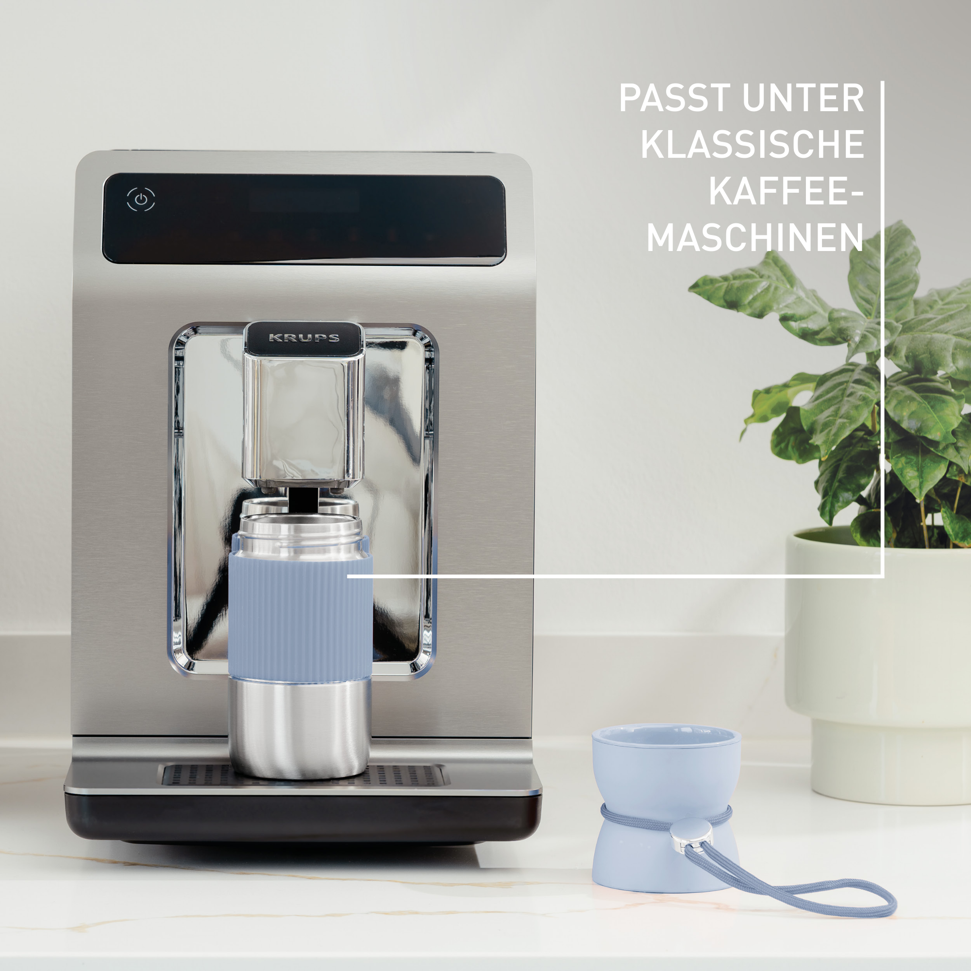 Eine Kaffeemaschine aus Edelstahl, eine blaue Tasse und eine Pflanze in einem weißen Topf sind im Bild.
