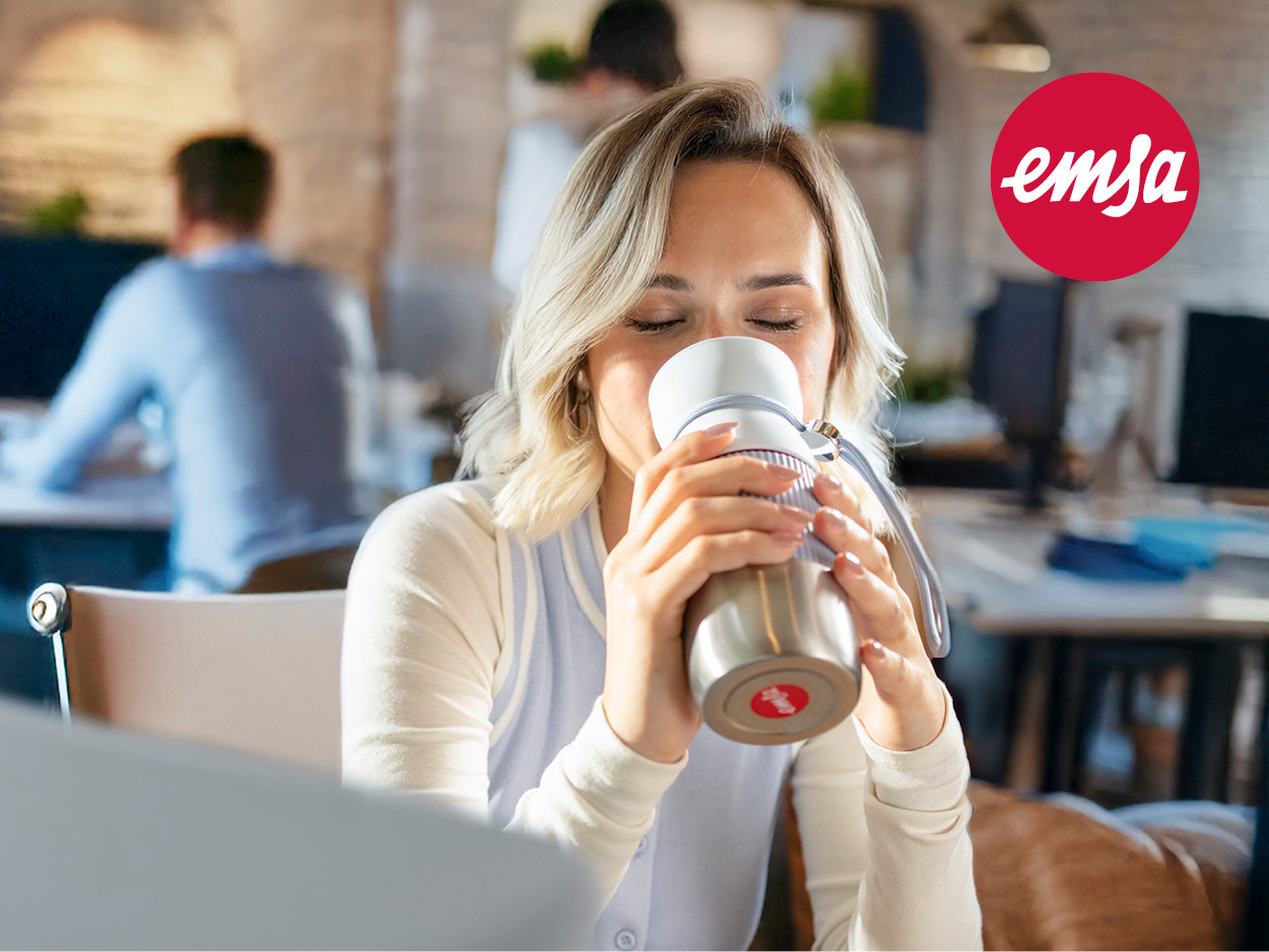 Eine Frau mit blonden Haaren trinkt im Büro aus einer silbernen Thermoskanne. Andere arbeiten im Hintergrund. Das Emsa-Logo ist sichtbar.