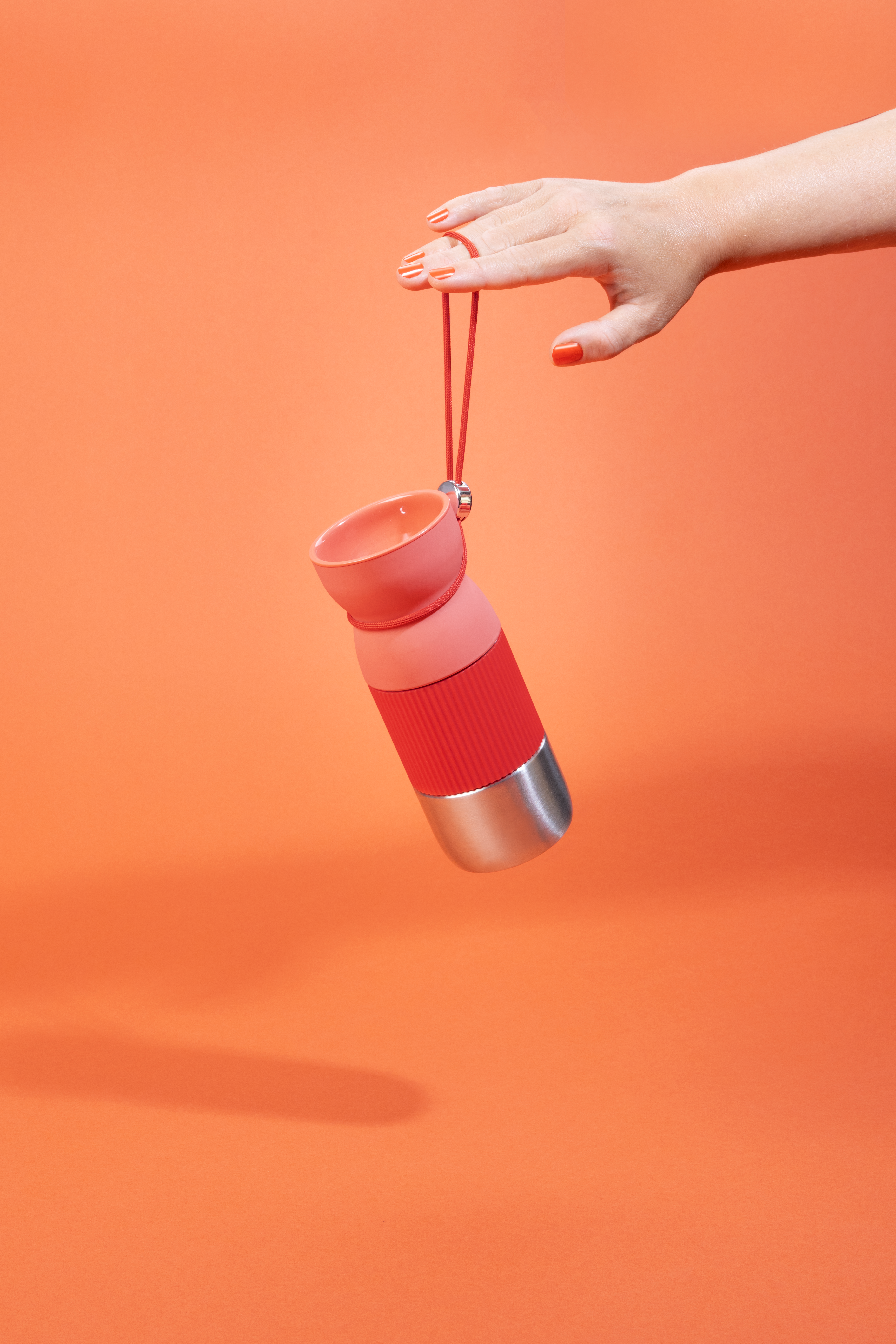 Eine Hand hält eine rot-rosa Wasserflasche vor orangefarbenem Hintergrund. Die Nägel sind rot lackiert.