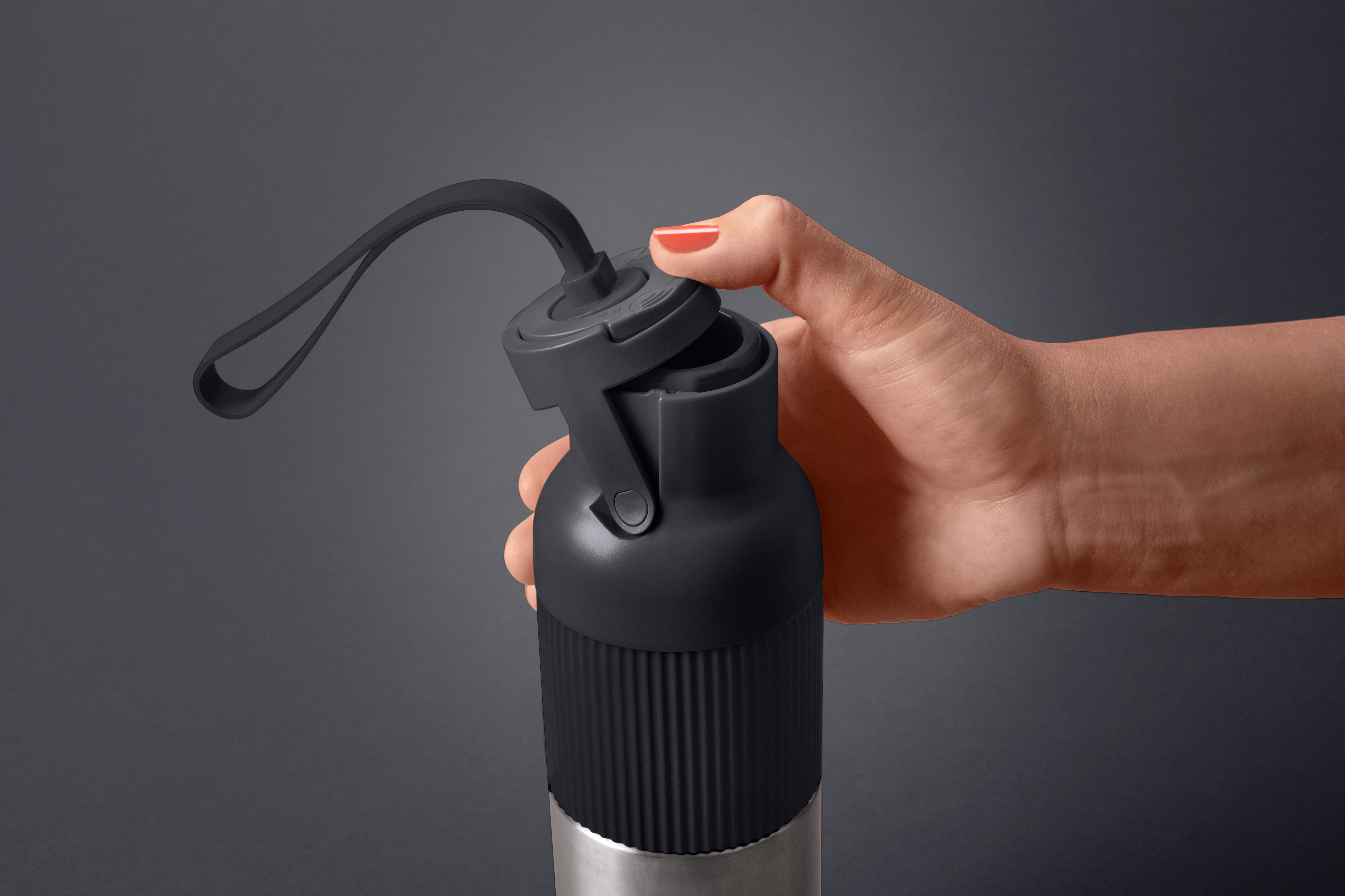 Eine Hand mit rotem Nagellack öffnet eine schwarze Wasserflasche vor grauem Hintergrund. Die Flasche hat einen silbernen Boden.