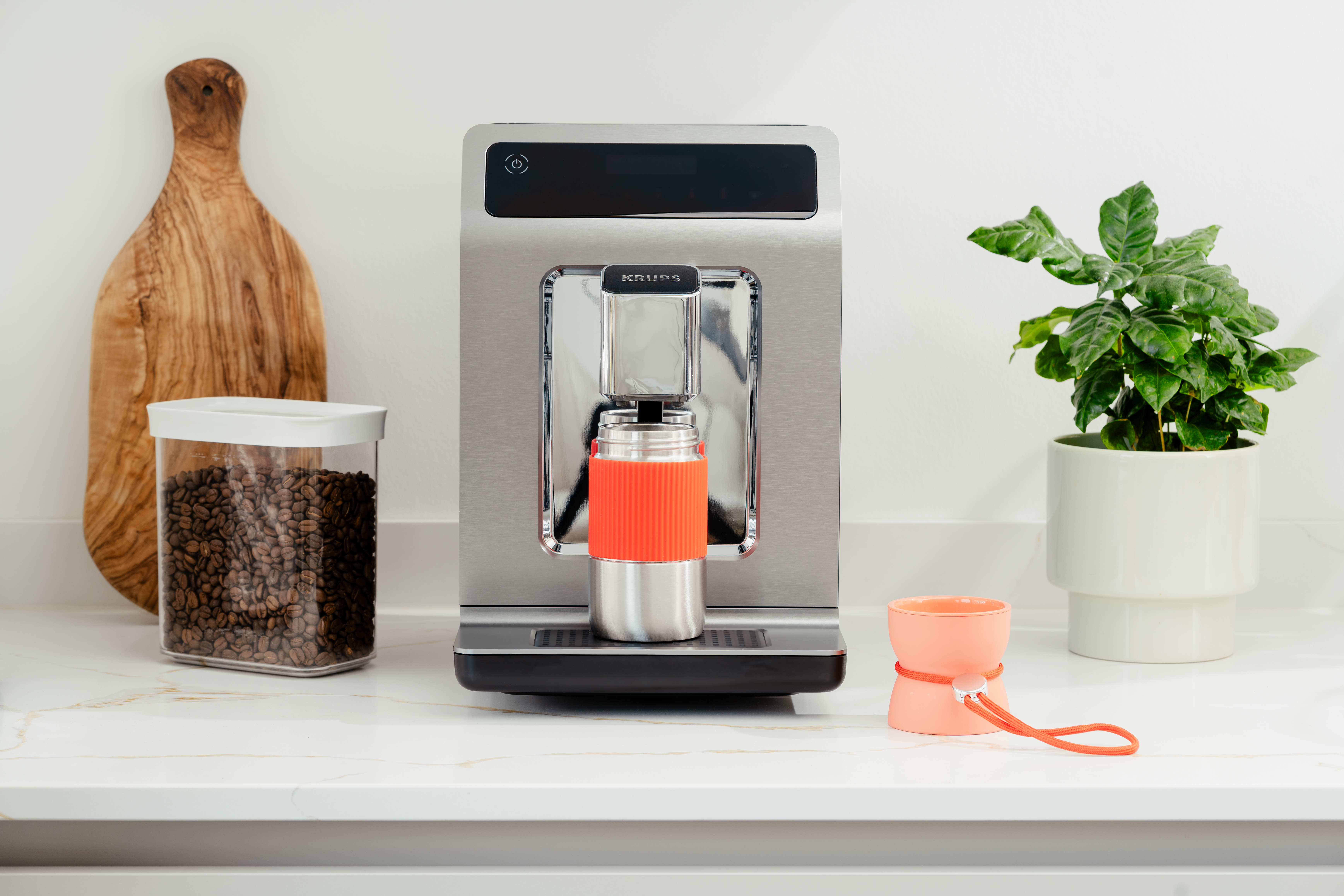 Eine Kaffeemaschine mit einer Tasse, einer Pflanze, einem Glas Bohnen und einer kleinen orangefarbenen Tasse auf einer weißen Arbeitsplatte.