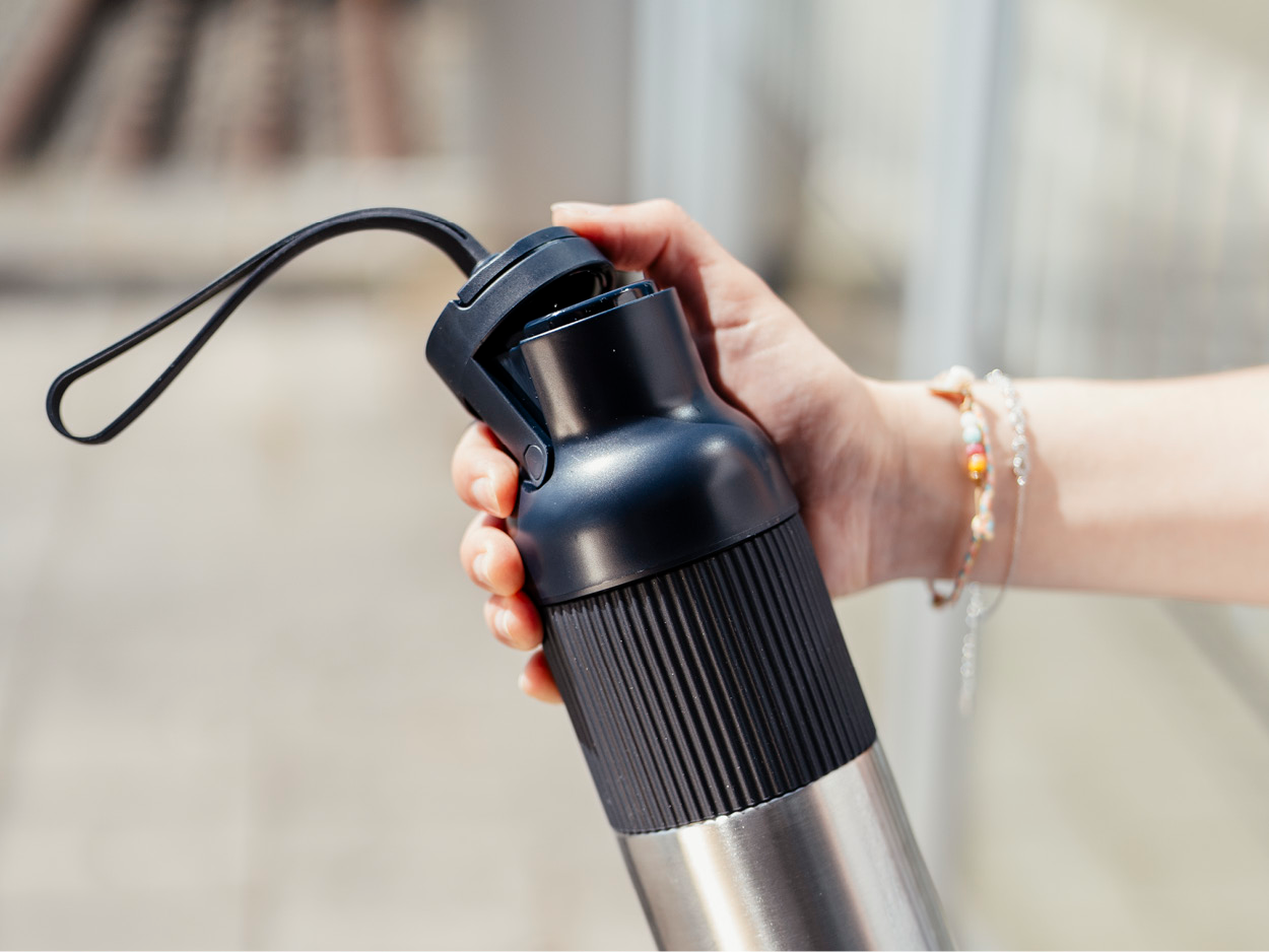 Eine Hand hält eine schwarz-silberne Wasserflasche mit Schlaufe. Schmuck verziert das Handgelenk. Der Hintergrund ist unscharf.