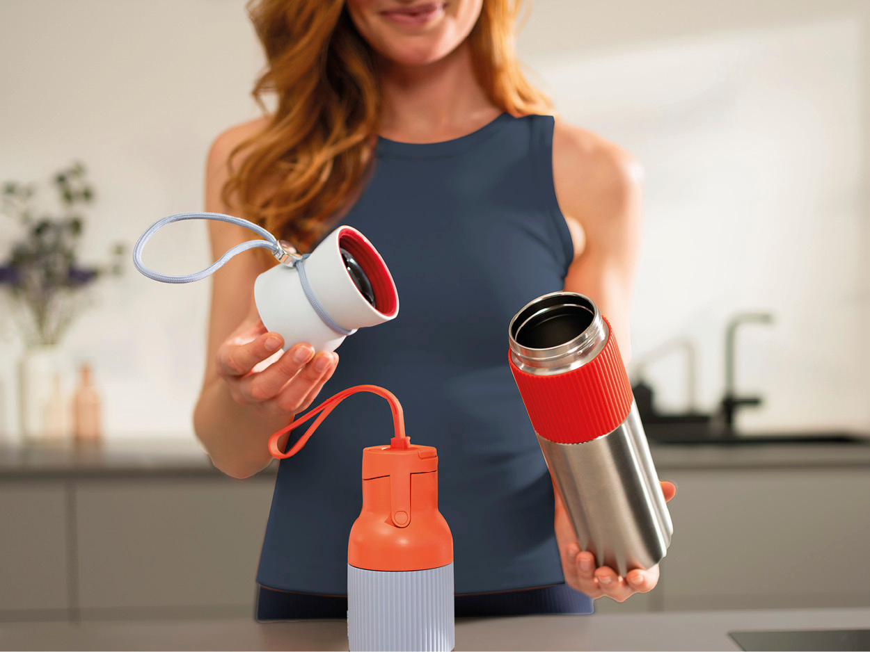 Frau hält Wasserflaschen: weiß und roter Deckel, orange und grau, und eine silberne Flasche mit rotem Band. Küchenhintergrund.