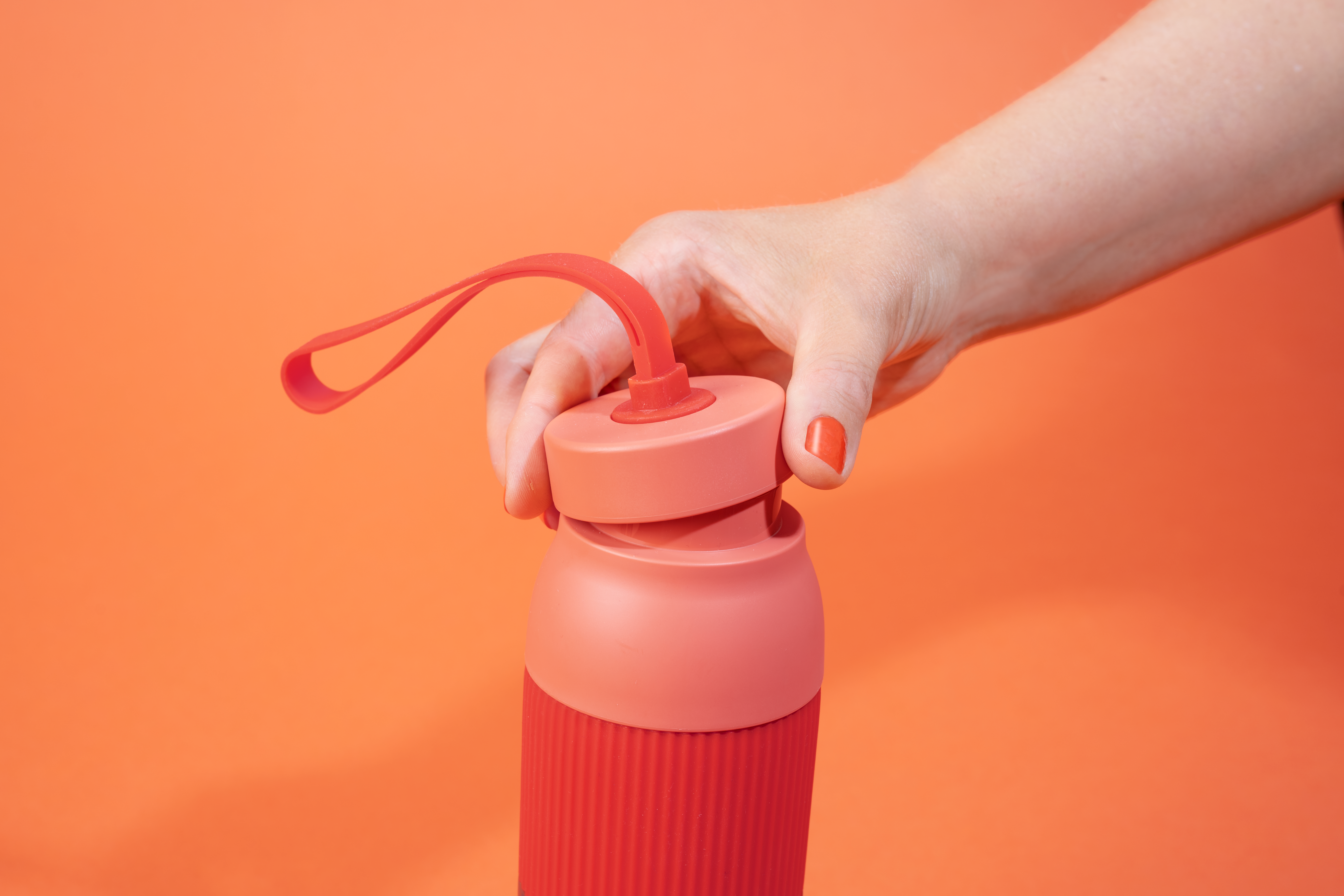 Eine Hand hält eine rote Wasserflasche. Der Hintergrund ist orange. Der Nagellack passt zur Flasche und zum Griff.