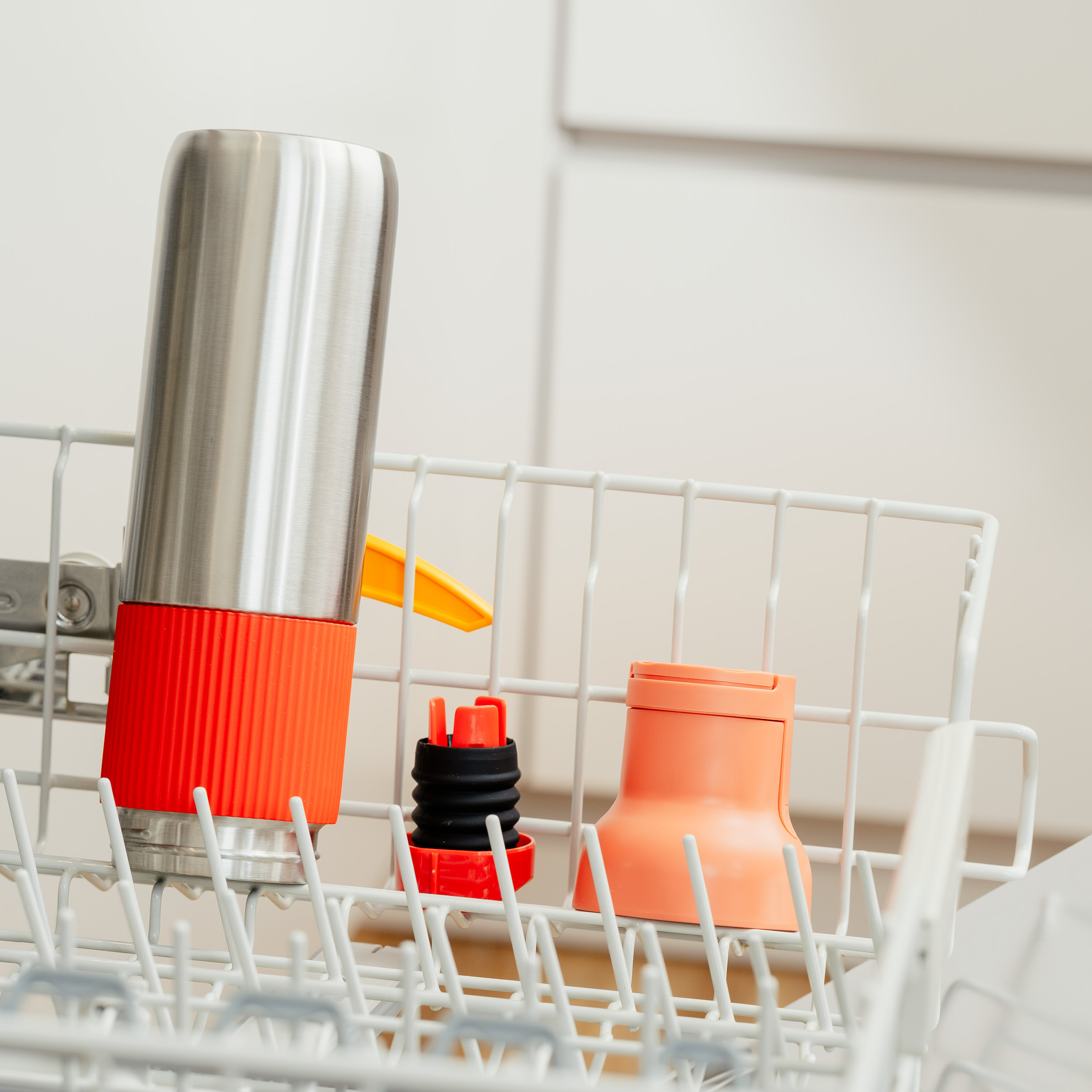 Geschirrspüler mit Edelstahlflasche, orangefarbener Tasse und schwarzem Deckel. Sie stehen in einem weißen Gestell. Hintergrund ist weiß.