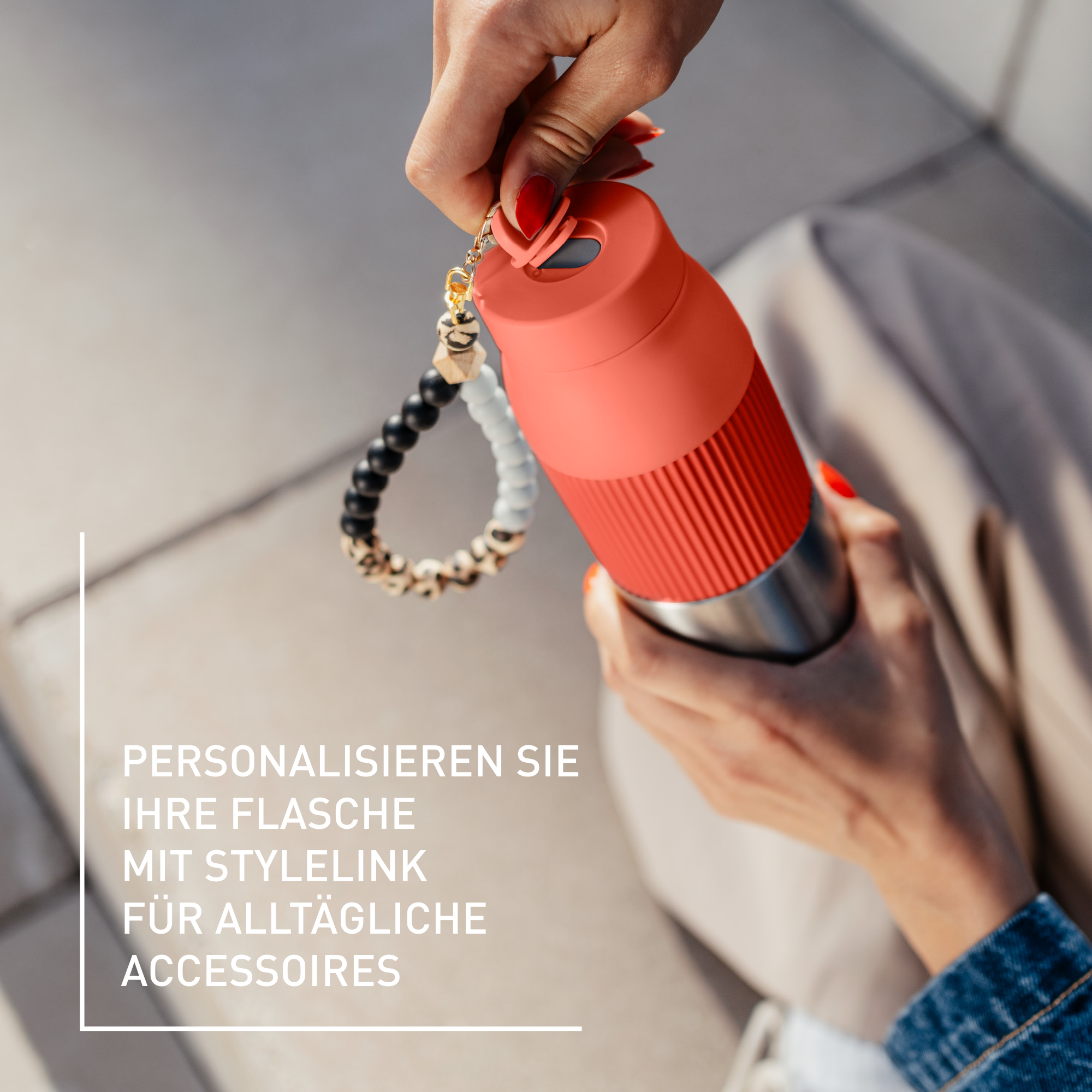 Eine Frau hält eine korallenfarbene Wasserflasche mit einem Perlenarmband. Rote Nägel sind sichtbar. Text bewirbt das Produkt.