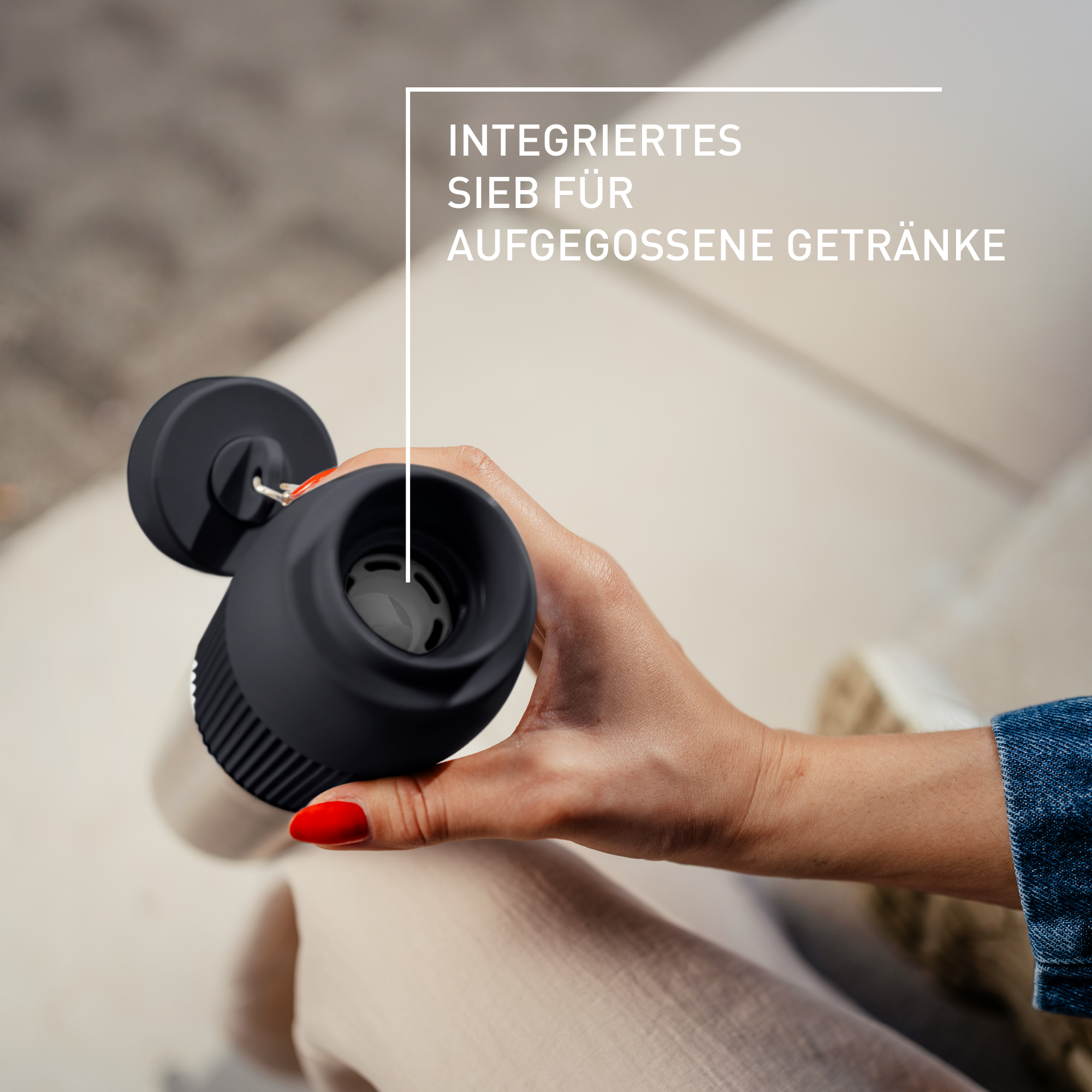 Eine Hand, die eine schwarze Tasse mit Filter hält. Roter Nagellack. Der Text INTEGRATED SIEB FÜR AUFGEGOSSENE GETRÄNKE ist sichtbar.