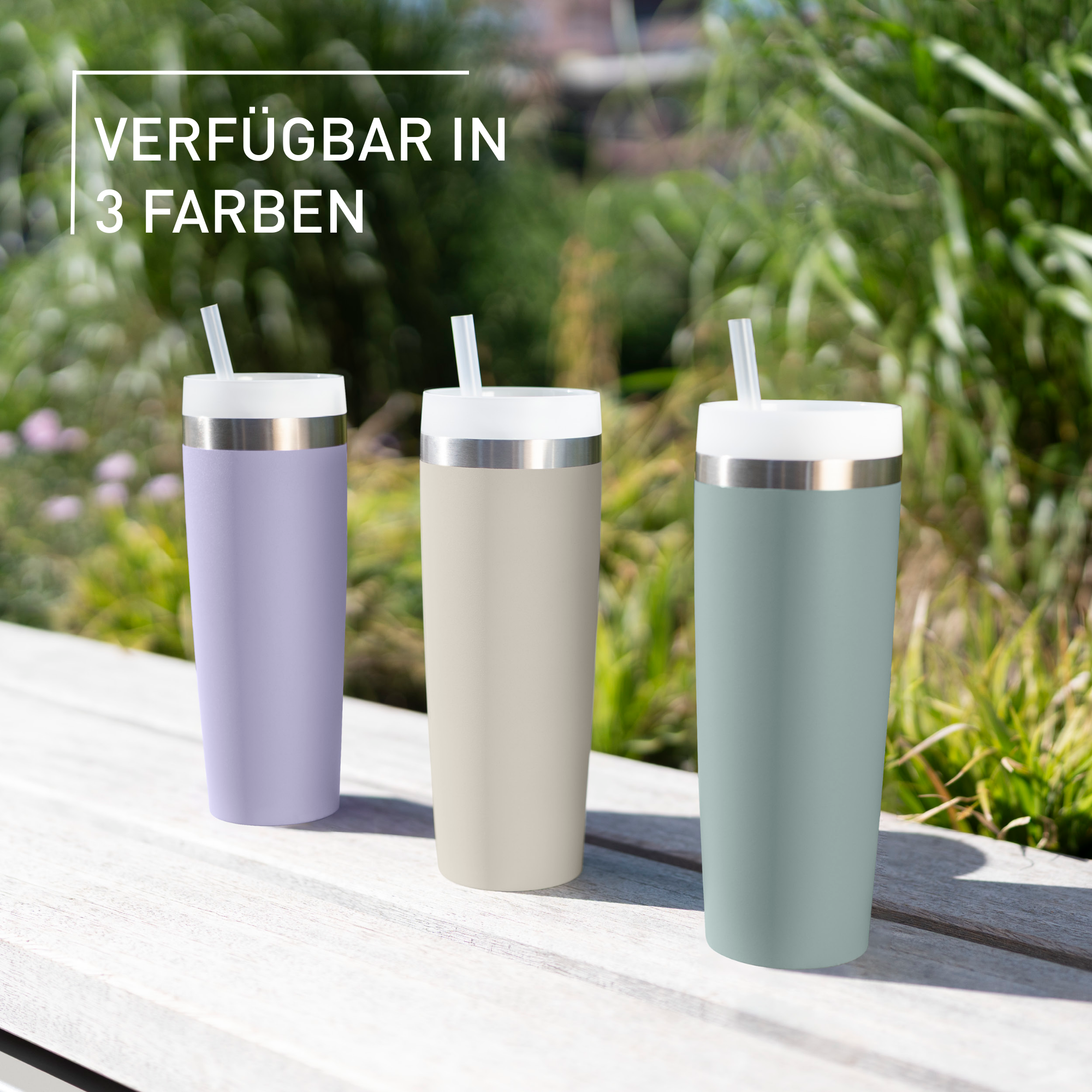 Drei Becher, lila, beige und grün, mit Strohhalmen, auf einer Holzbank, mit Grün im Hintergrund.