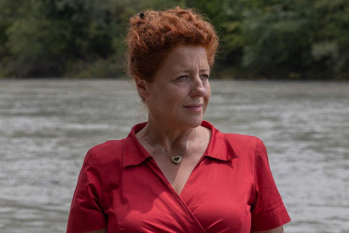 Eine Frau mit roten Haaren und rotem Hemd steht am Fluss. Sie trägt eine Herzanhängerkette und blickt zur Seite.