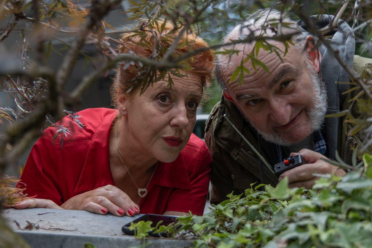 Zwei Personen schauen über eine Mauer. Eine Frau in Rot und ein Mann mit grauem Bart halten ein Gerät. Pflanzen im Vordergrund.