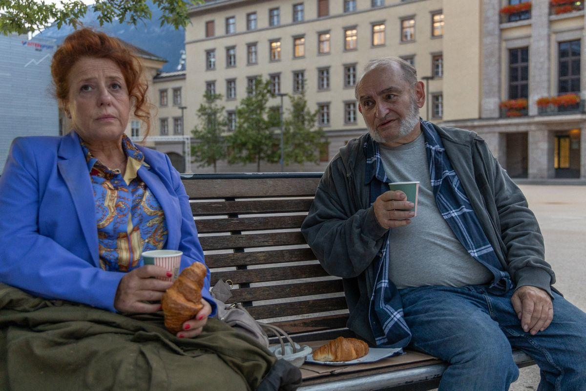 Zwei Personen sitzen auf einer Bank und halten Tassen und Croissants. Im Hintergrund sind ein Gebäude und Bäume.