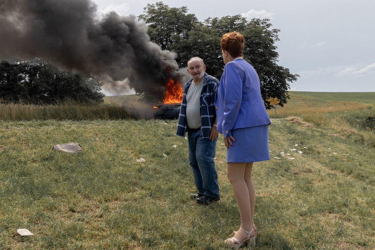 Zwei Personen beobachten ein brennendes Auto auf einem Feld. Schwarzer Rauch steigt auf. Eine Person trägt Blau, die andere Jeans und ein kariertes Hemd.