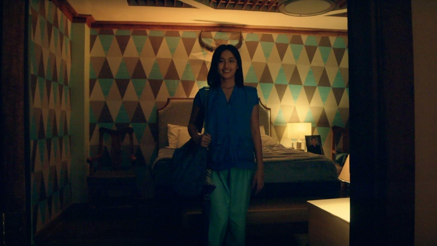 Eine lächelnde Frau in einem Raum. Sie hält eine Tasche und trägt ein blaues Oberteil und eine grüne Hose. Hinter ihr sind ein Bett und eine gemusterte Wand.