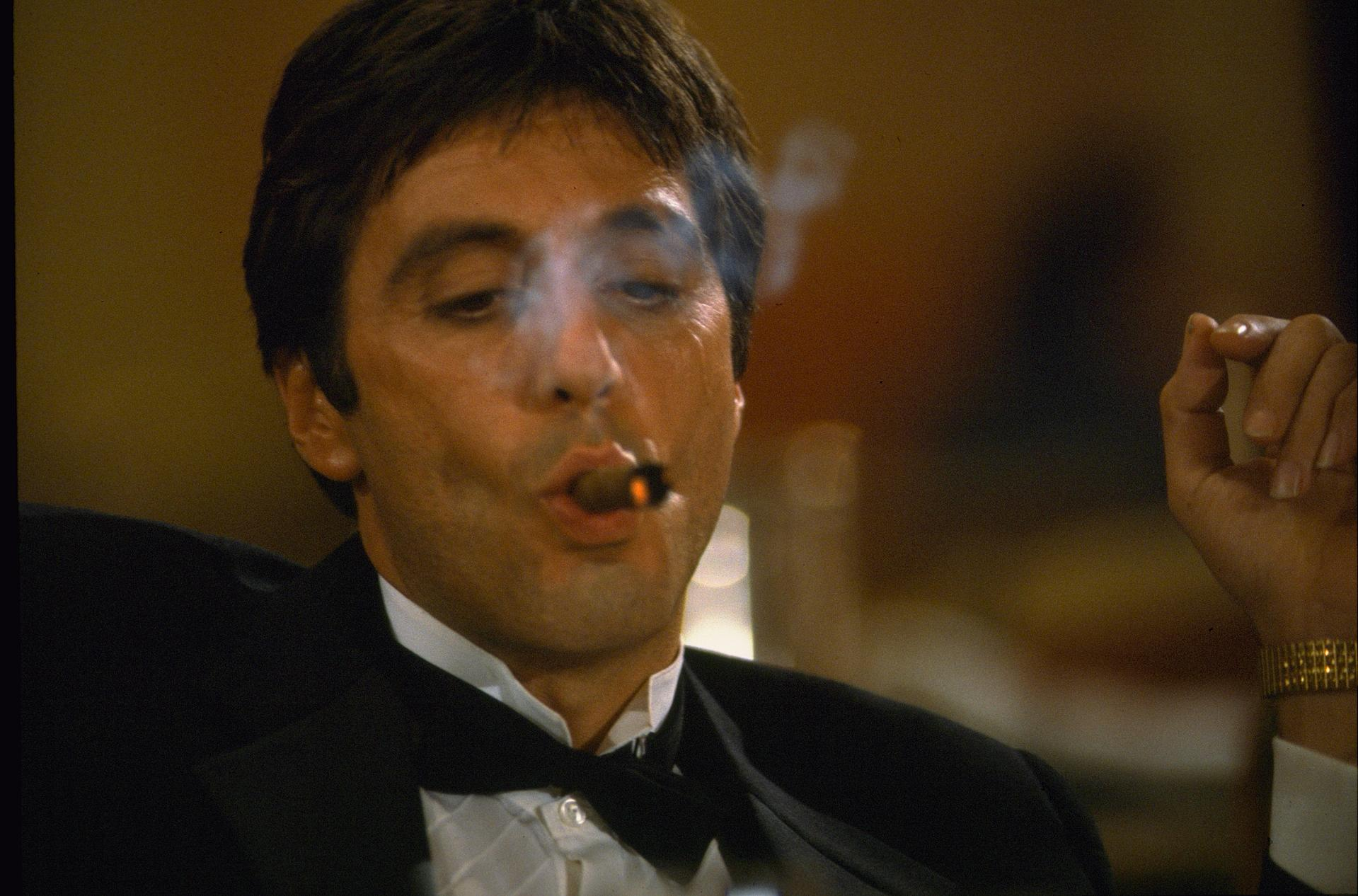 Mann im Smoking raucht eine Zigarre und stößt Rauch aus. Goldene Uhr sichtbar. Dunkler Hintergrund mit warmen Tönen.