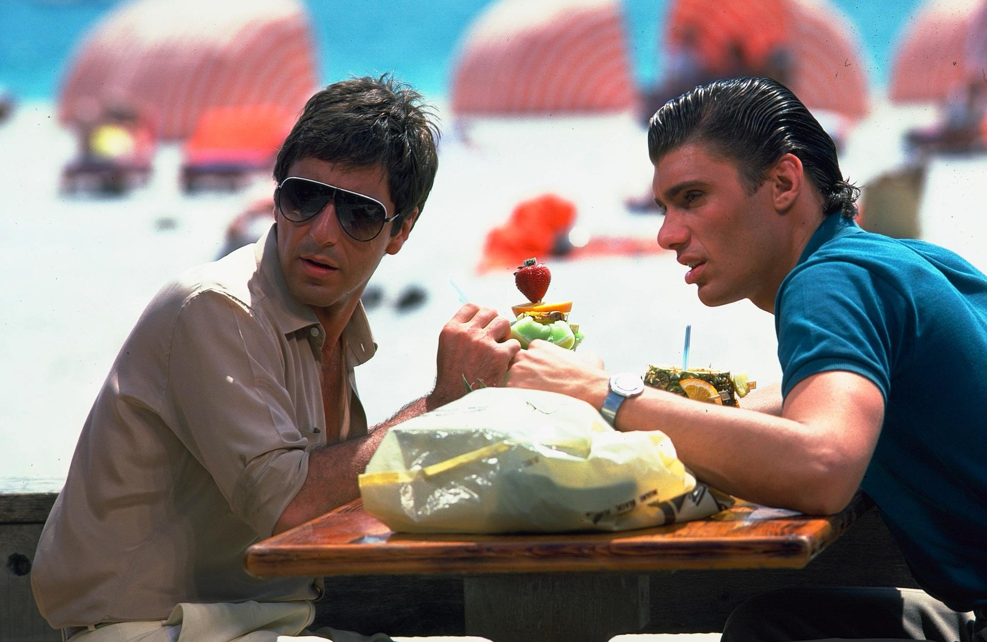 Zwei Männer sitzen an einem Tisch. Einer trägt eine Sonnenbrille, der andere ein blaues Hemd. Ein Obstspieß steht zwischen ihnen. Hintergrund: Strandszene.
