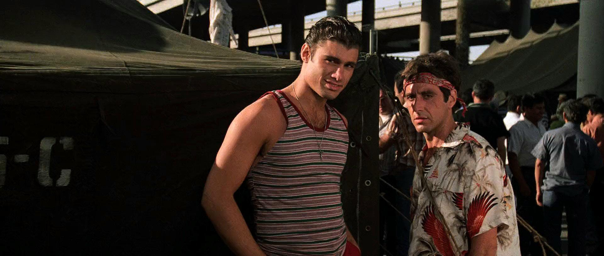 Zwei Männer stehen draußen. Einer trägt ein gestreiftes Tanktop, der andere ein Blumenhemd und ein Bandana. Im Hintergrund sind Leute.