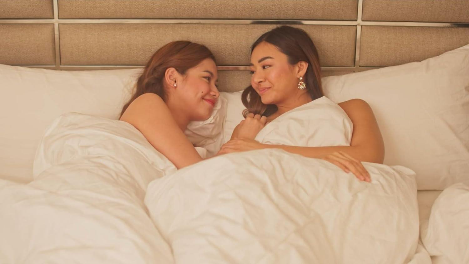 Zwei Frauen im Bett, die sich anlächeln. Sie sind von weißen Laken bedeckt. Der Hintergrund ist ein beiges Kopfteil.