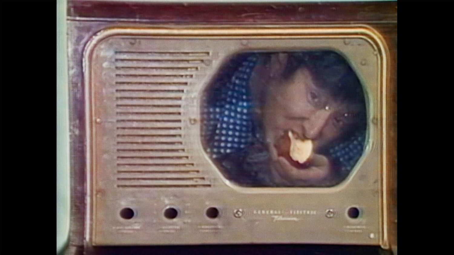 Eine Person in einem blau gepunkteten Hemd isst etwas. Die Person befindet sich in einem alten General Electric Fernseher.