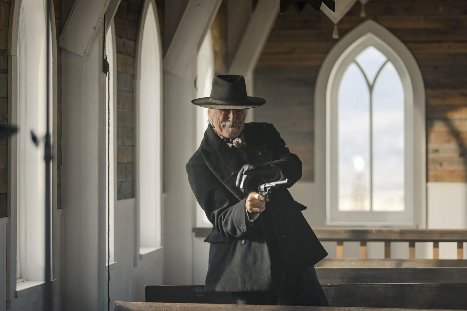 Ein älterer Mann mit schwarzem Hut und Mantel zielt mit einer Pistole. Er steht in einem kirchenähnlichen Gebäude.