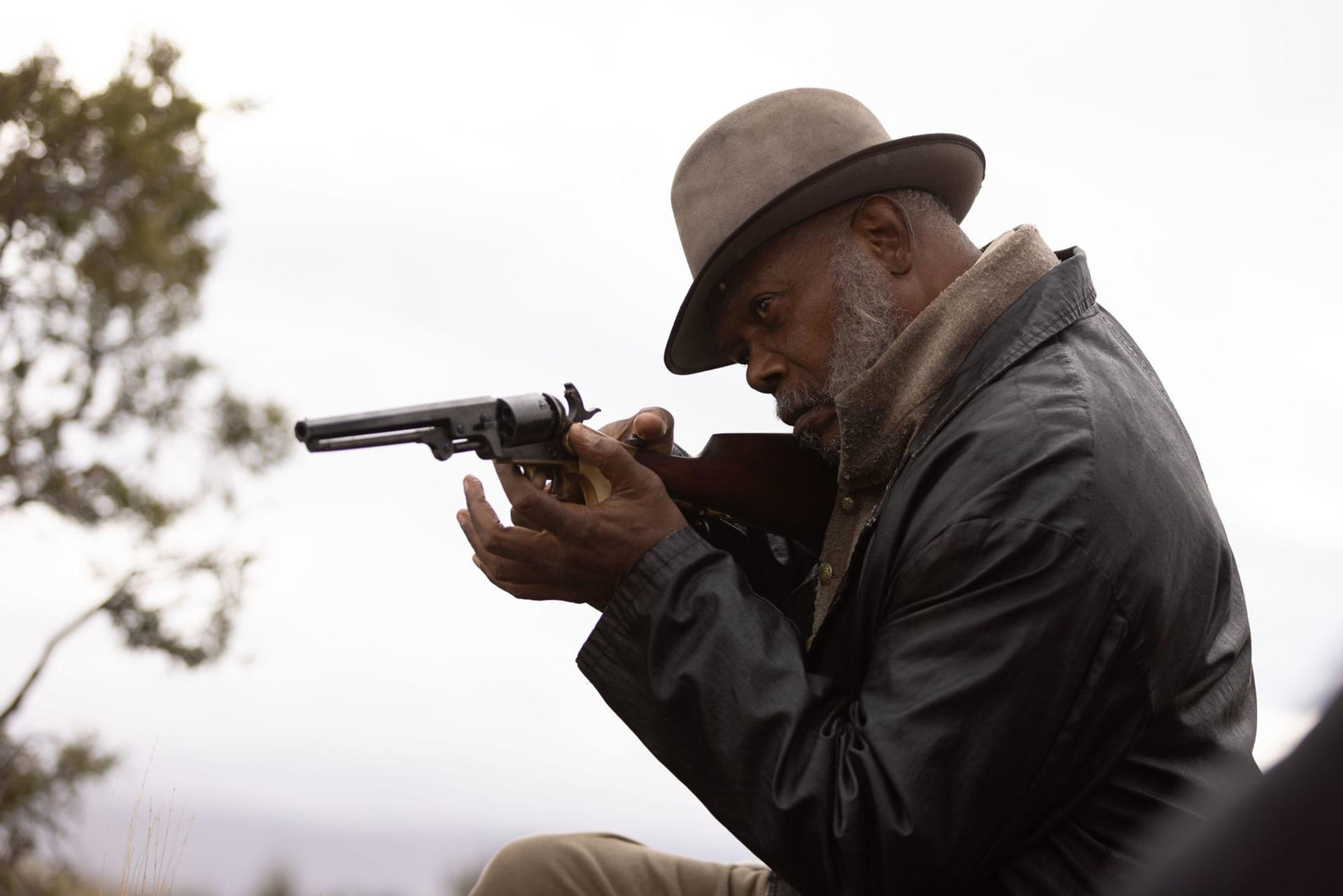 Ein älterer schwarzer Mann zielt mit einem Revolver. Er trägt einen Hut und einen Mantel. Der Hintergrund ist eine verschwommene Landschaft und Himmel.