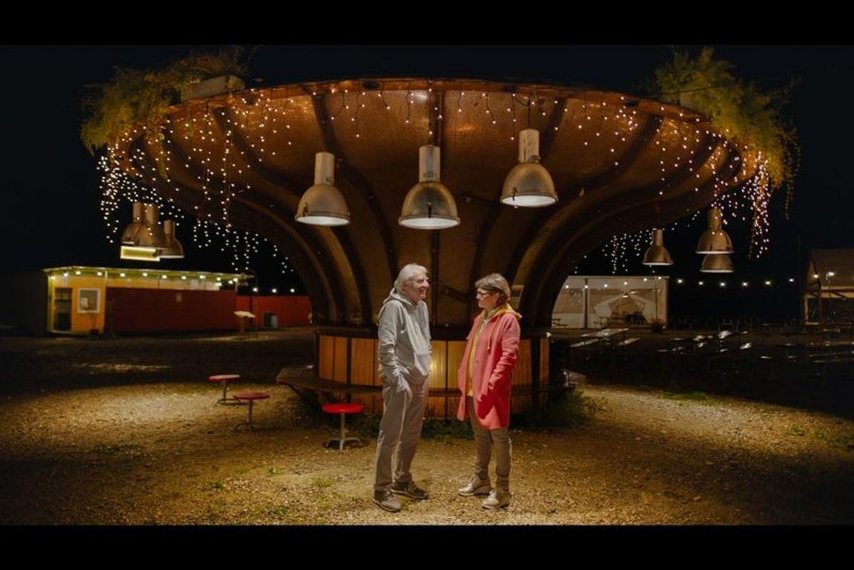 Zwei Personen stehen unter einer großen Struktur mit hängenden Lichtern. Gebäude sind im Hintergrund zu sehen, Nachtszene.