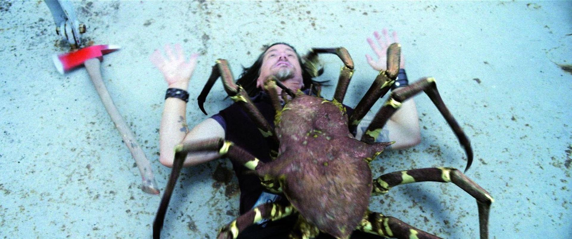 Ein Mann liegt auf einer Oberfläche, eine große Spinne auf ihm, eine Axt darüber. Der Mann hebt die Hände.
