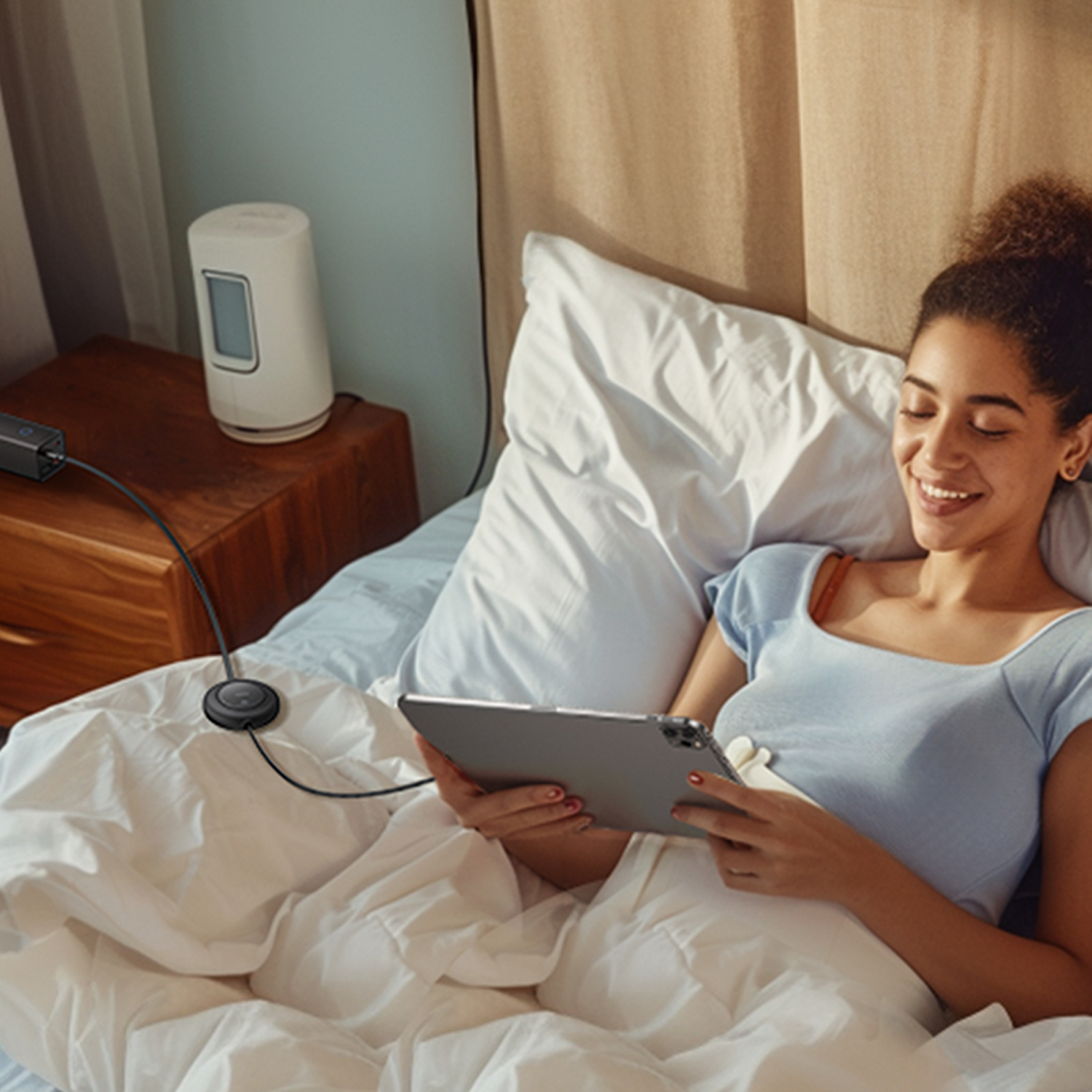 Mujer en la cama sonríe usando una tableta. Junto a ella, una mesita de noche con un dispositivo de carga.