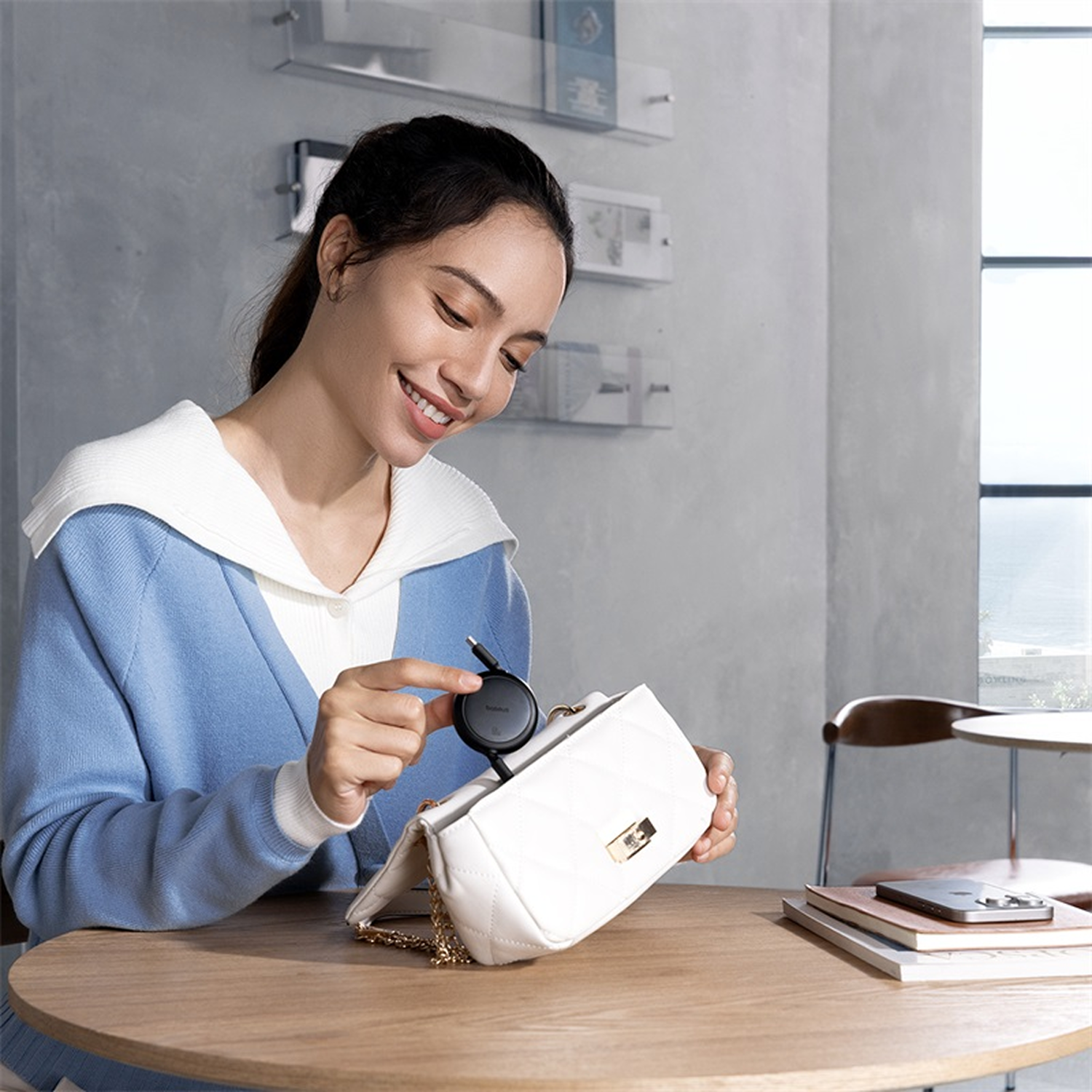 Mujer sonríe, sosteniendo un dispositivo negro de un bolso blanco. Mesa con teléfono y libros. Fondo: ventana y decoración mural.