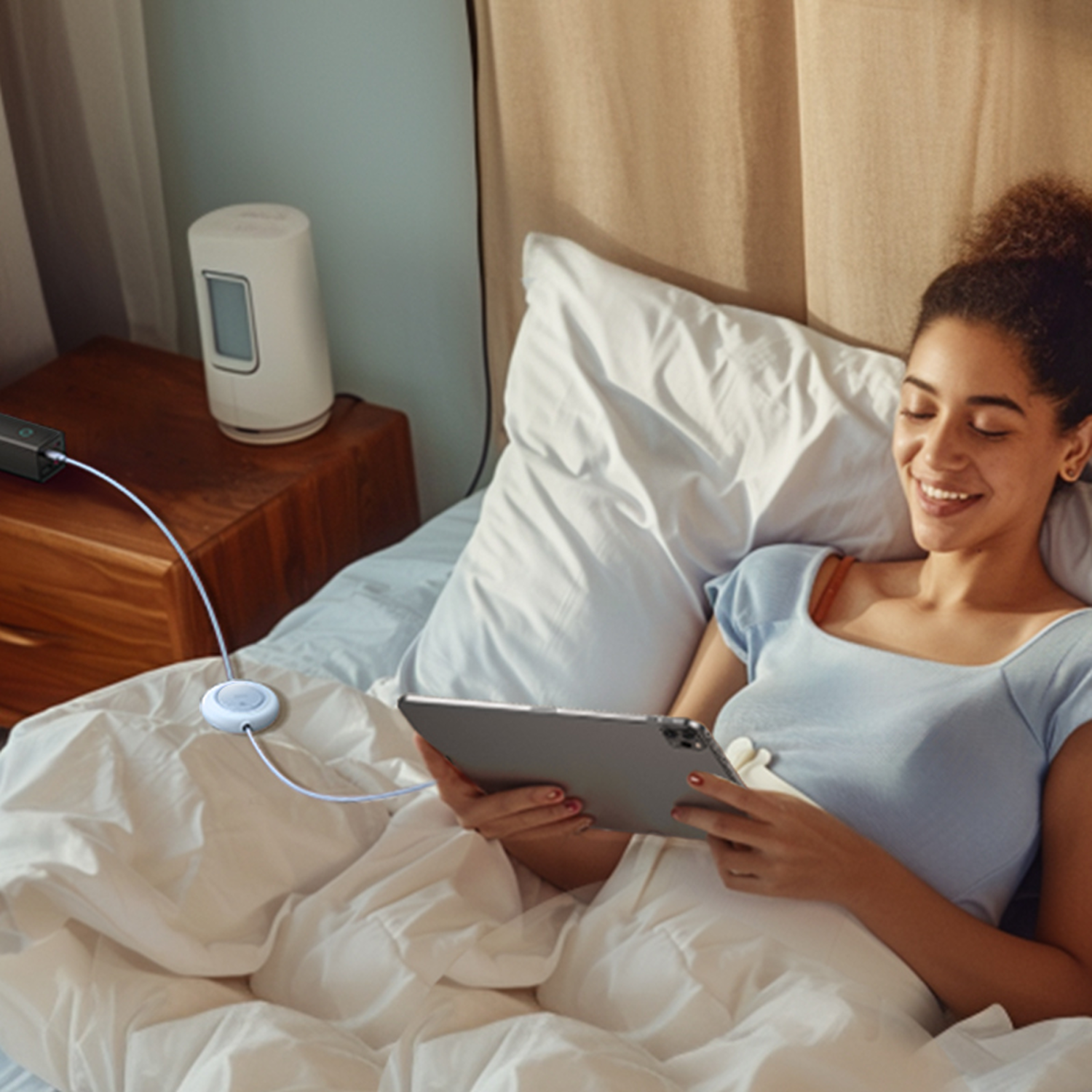 Mujer en la cama, sonriendo, sosteniendo una tableta. Se ve una mesita de noche con un dispositivo y un cargador.