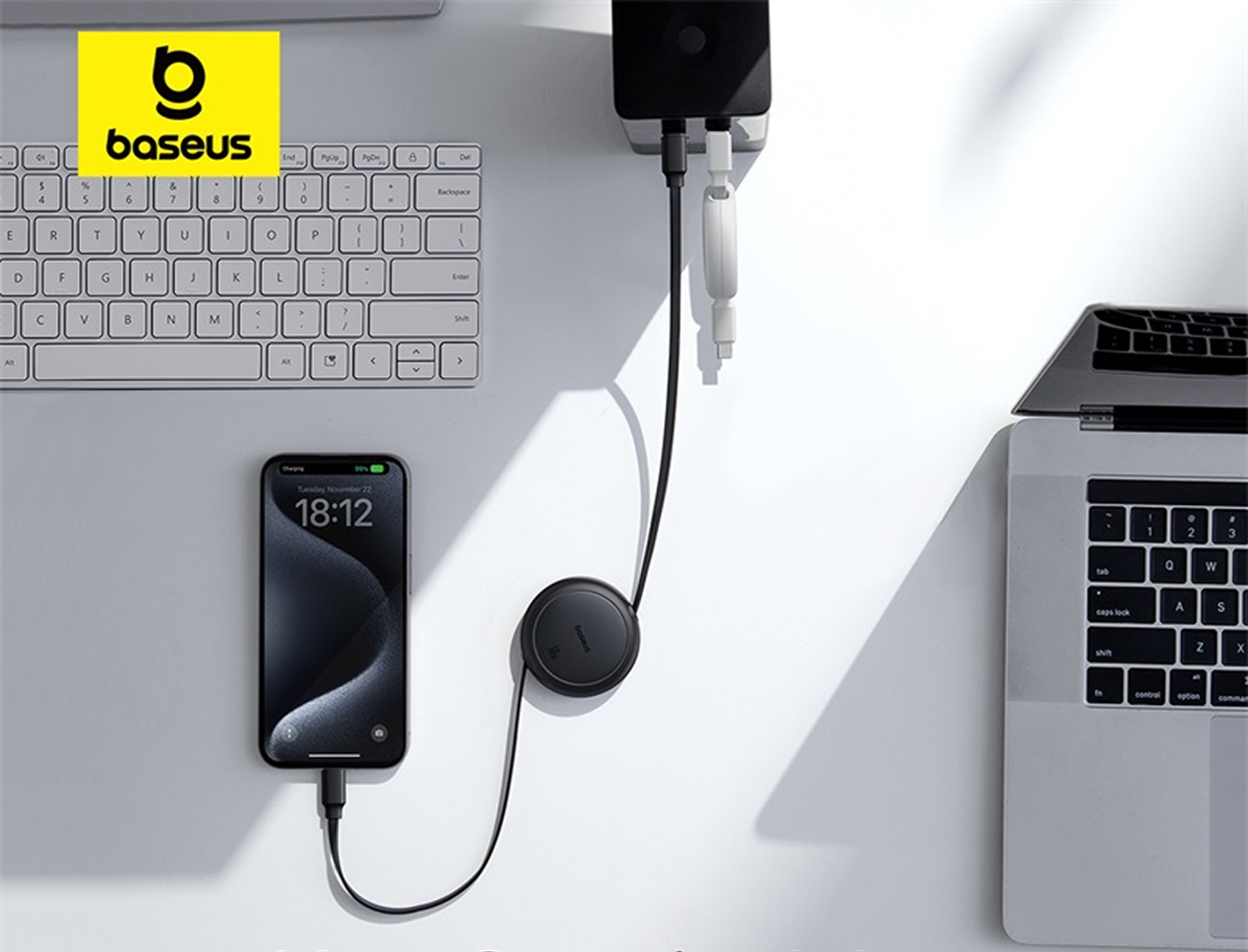 Un teléfono cargando en un escritorio blanco, junto a un portátil y un teclado. Un concentrador de carga negro está conectado a la pared.