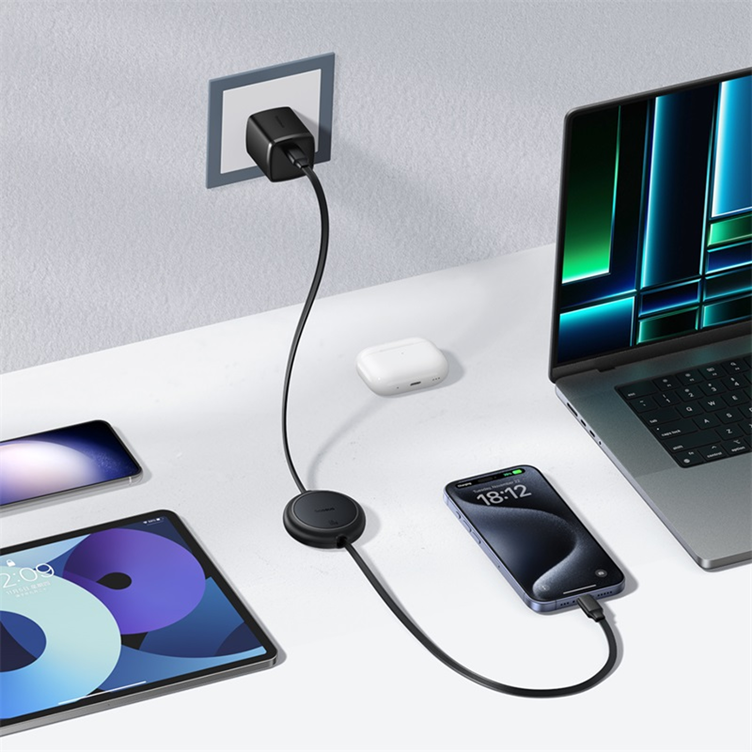 Un cargador alimenta varios dispositivos: portátil, teléfono, tableta y un teléfono, y un estuche de carga. Todos los dispositivos están sobre una superficie blanca.