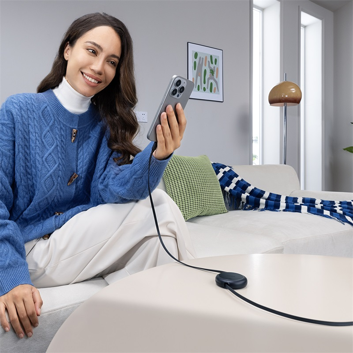 Mujer en suéter azul usa teléfono conectado a una plataforma de carga en la mesa. Sonríe, sentada en un sofá.