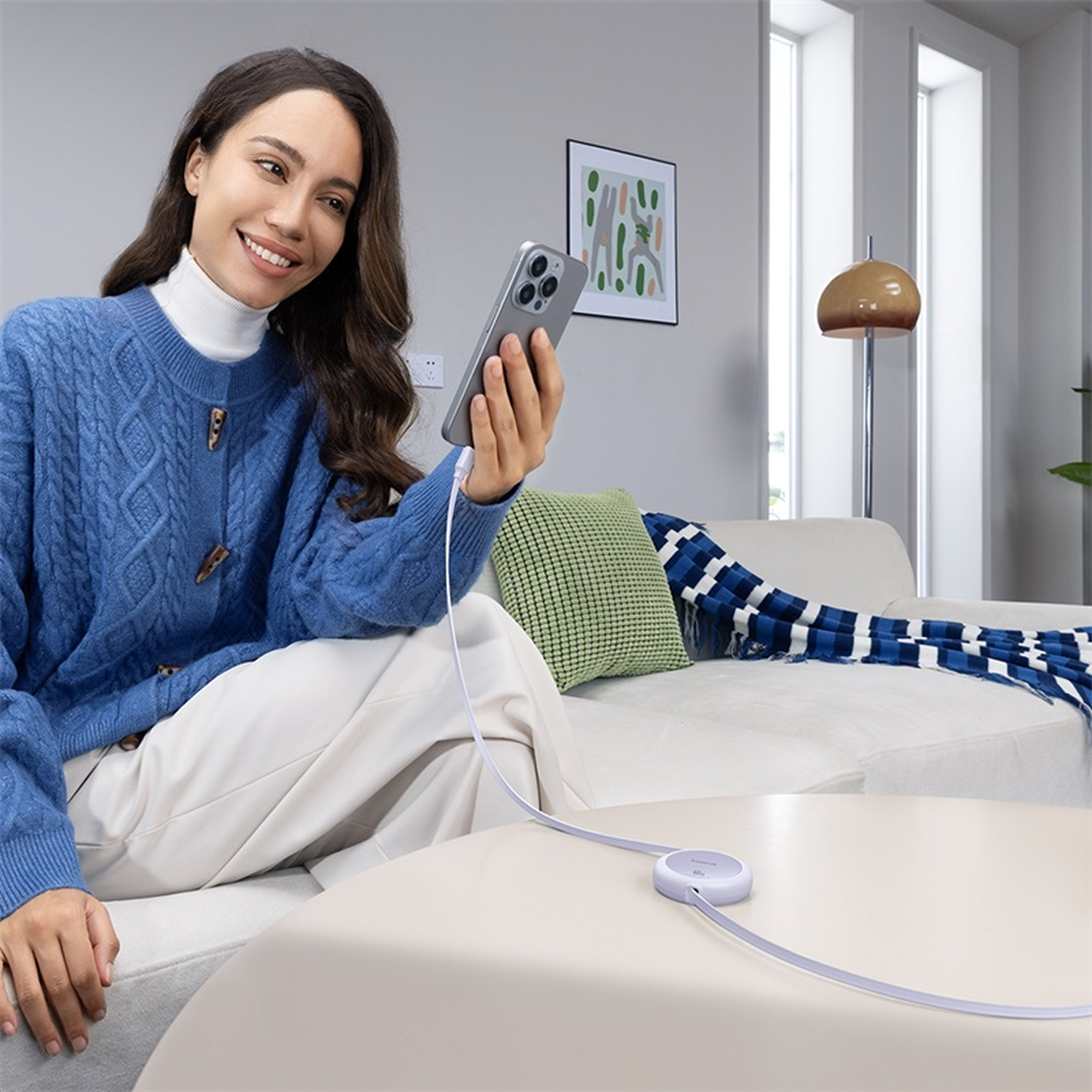 Mujer en suéter azul usa teléfono, cargando en mesa. Sofá con almohadas y manta en el fondo, lámpara y arte en la pared.