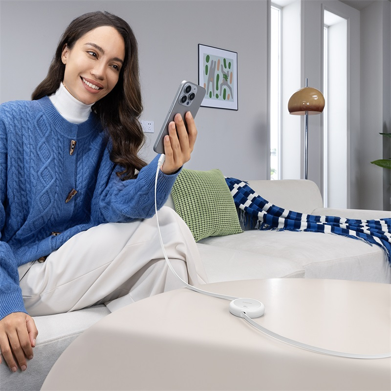 Mujer en suéter azul sosteniendo un teléfono, cargando. Sofá, mesa, lámpara y marco de imagen de fondo.