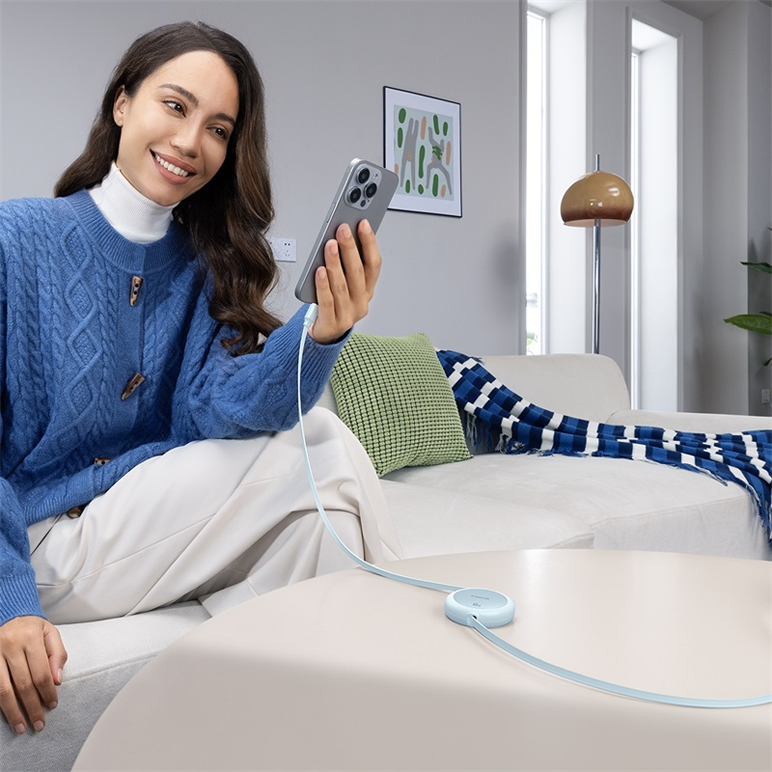 Mujer con suéter azul usa teléfono conectado a cargador en mesa. Sofá y lámpara al fondo.